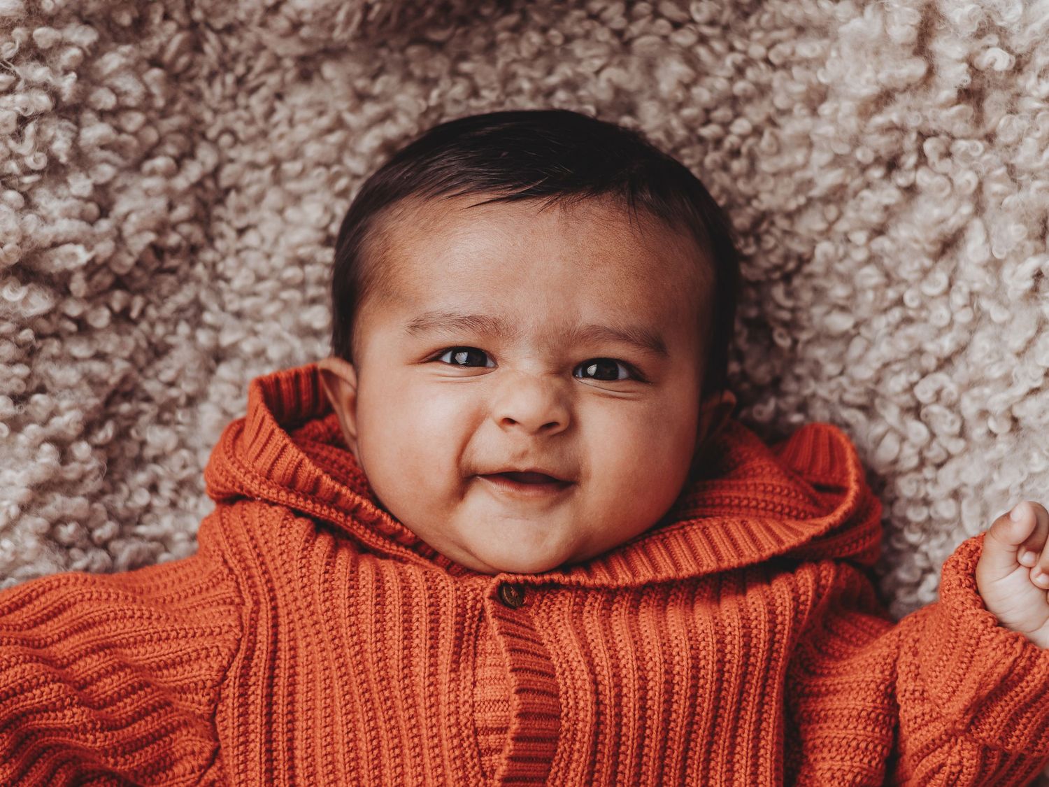 Baby wearing a cozy orange knit sweater smiles while lying on soft gray textured fabric.
