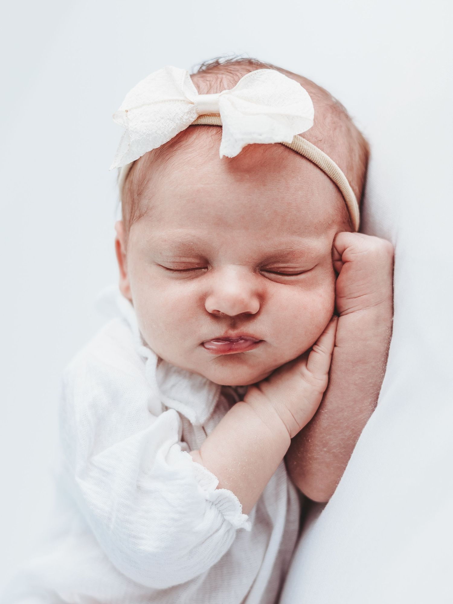 A sweet sleeping newborn baby wearing a white outfit and delicate bow headband rests peacefully in soft lighting.