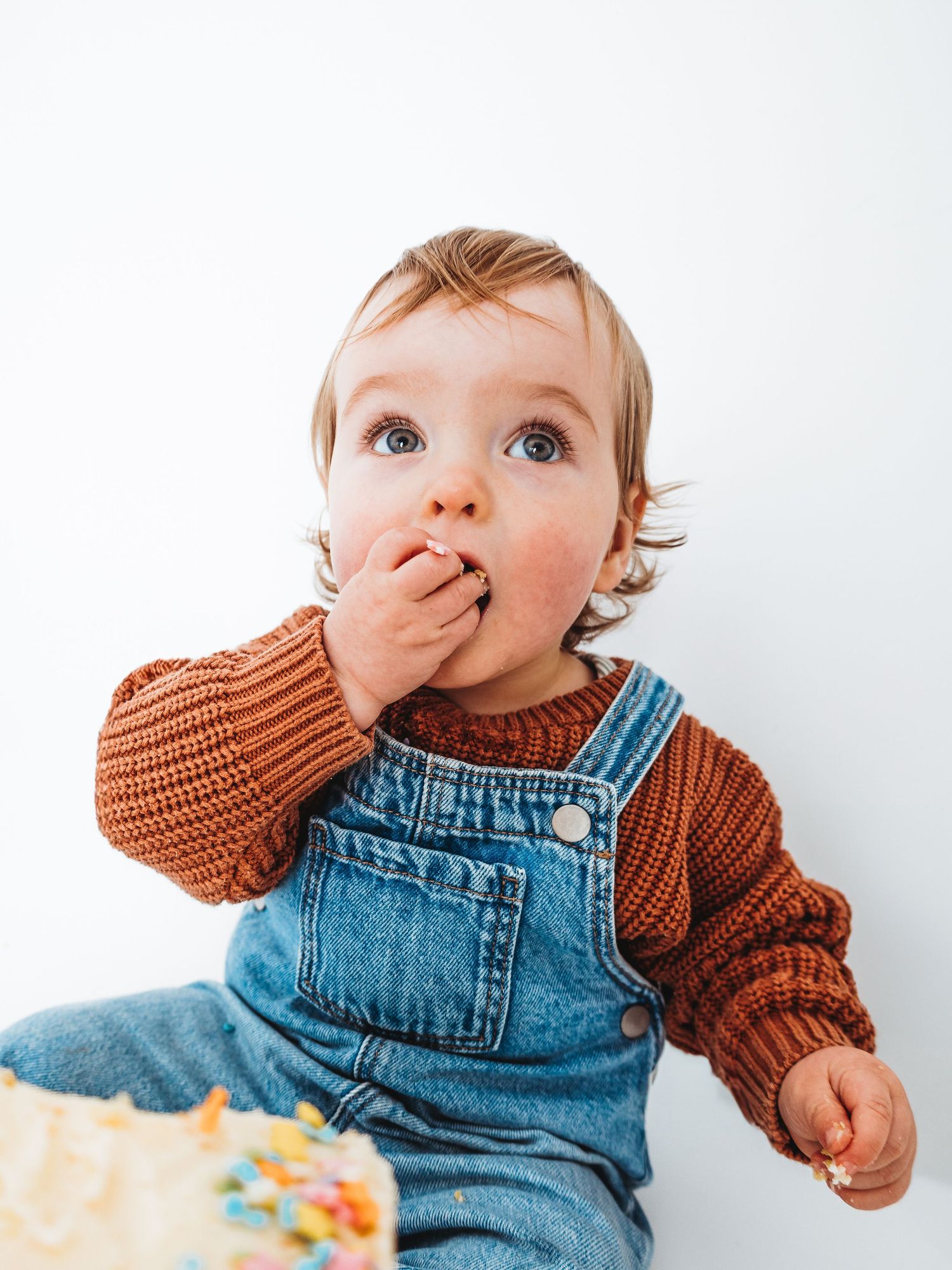 A cute baby in denim overalls and rust sweater eating birthday cake with their hand, smiling curiously.