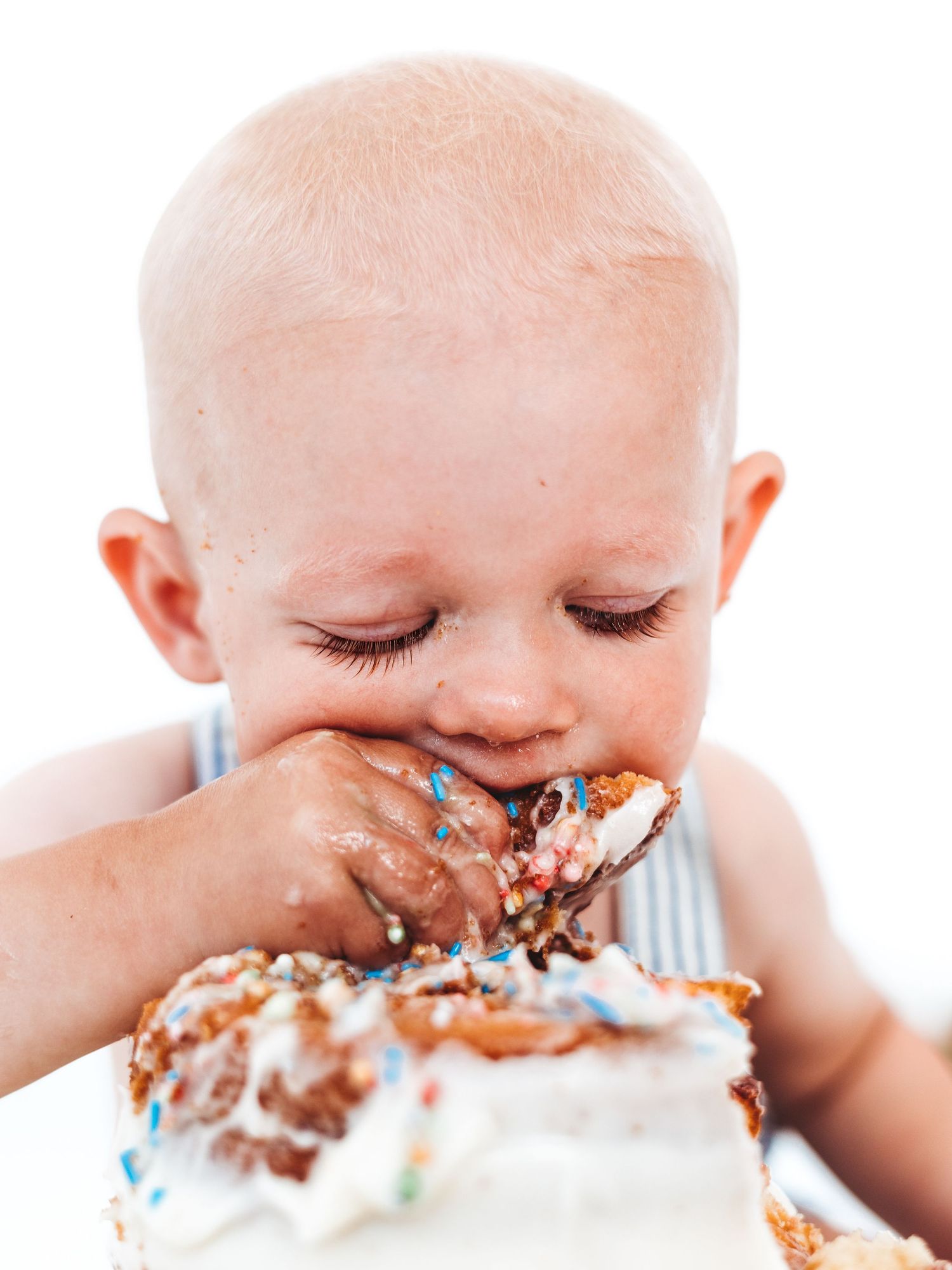 Adorable baby enjoying first birthday smash cake, covered in white frosting and colorful sprinkles.