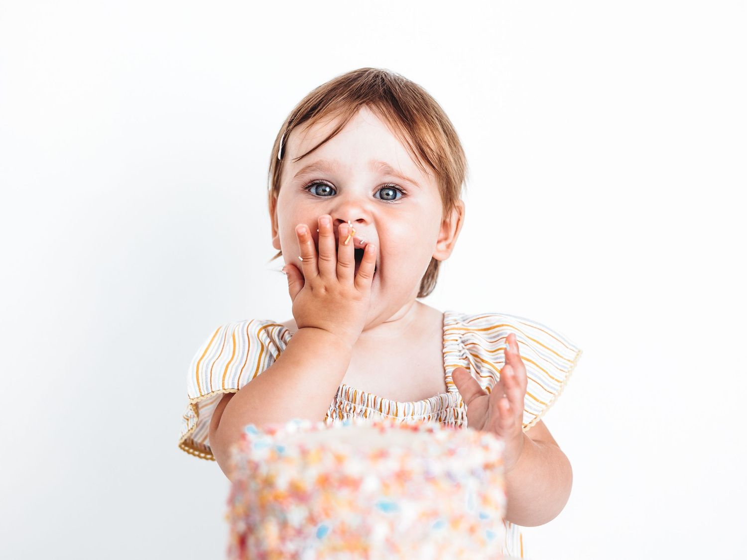 Cute toddler girl eating birthday cake with her hands, wearing a striped dress against a white background.