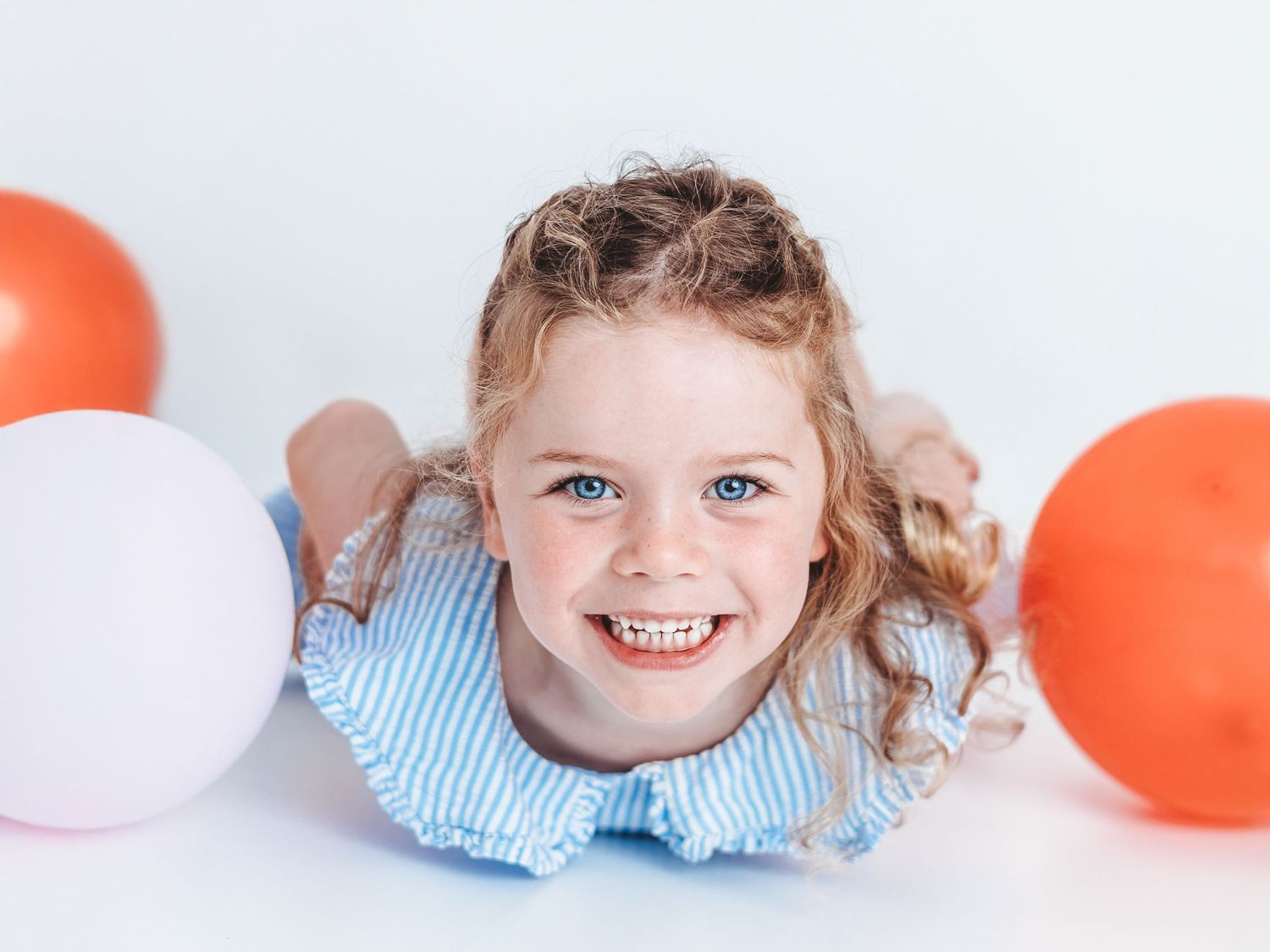 Smiling young girl with curly hair lying down surrounded by red and white balloons on a white background.