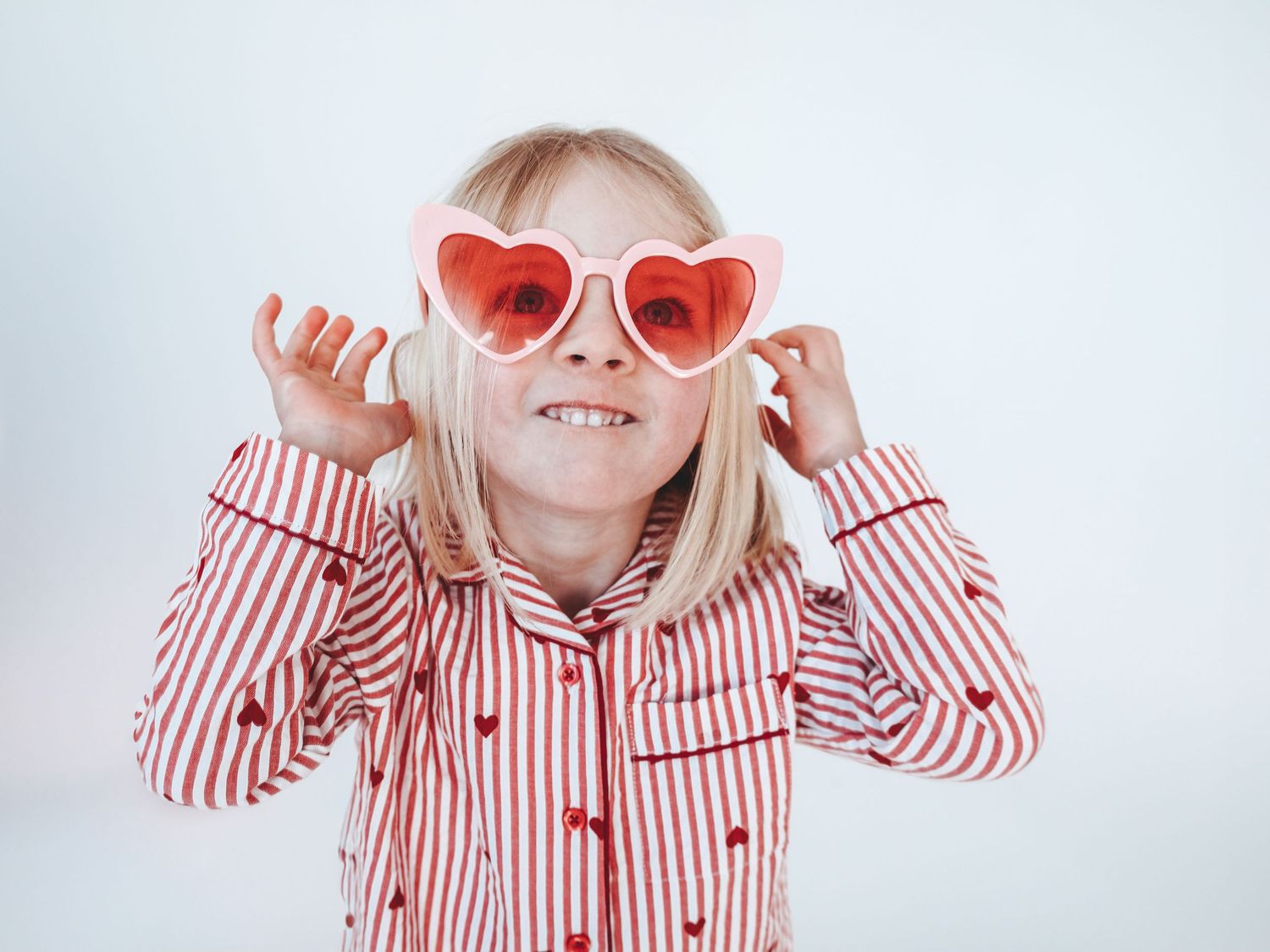 A smiling young girl wearing heart-shaped red sunglasses and a red striped shirt with heart prints on a white background.