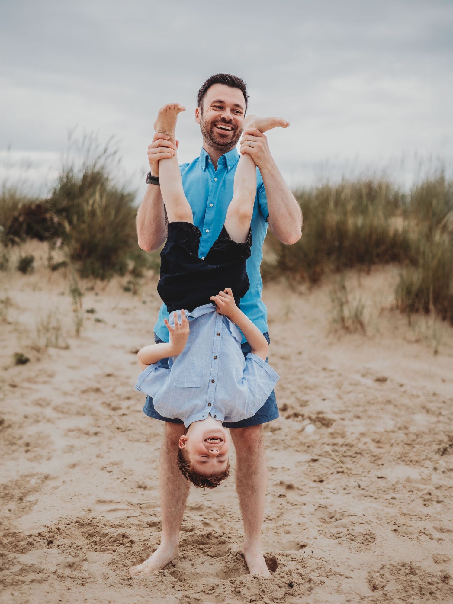 A laughing man holds a giggling child upside down on a sandy beach, both wearing blue shirts.