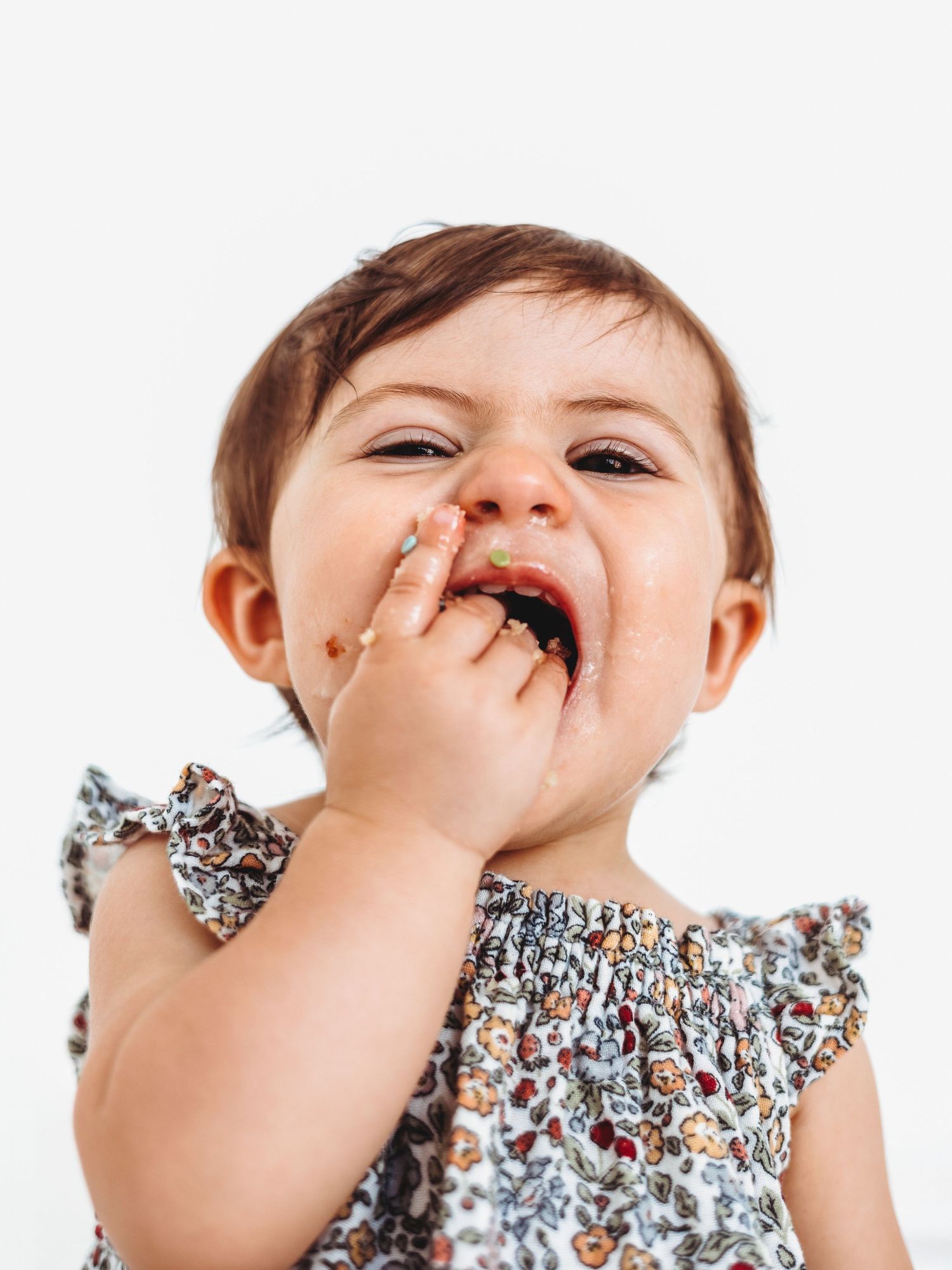 Adorable baby girl with food on her face, smiling and eating with her hand, wearing a floral dress.