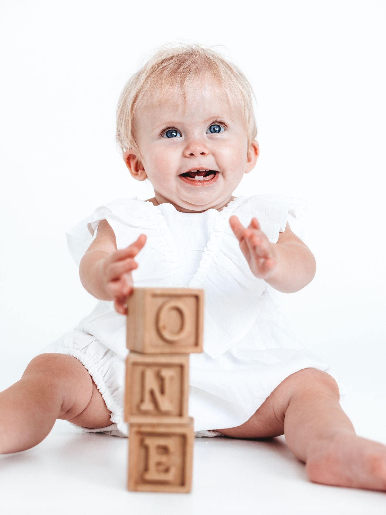 A smiling baby girl in a white dress sits with wooden alphabet blocks spelling ONE for her first birthday photo.