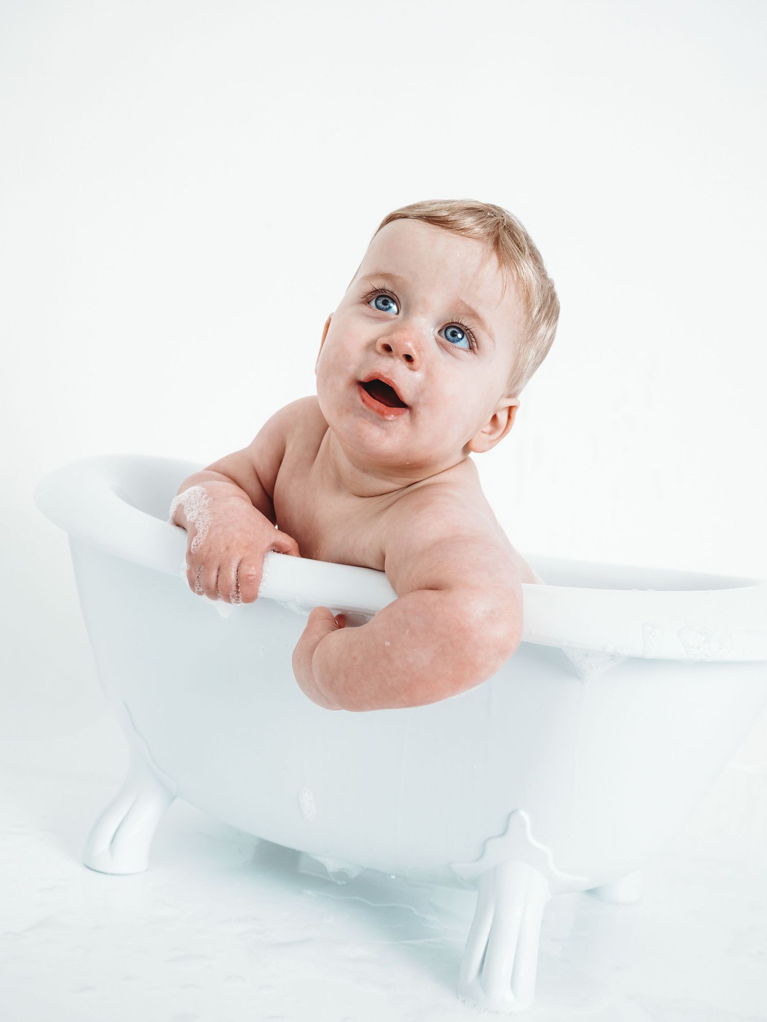 Cute baby with blue eyes sitting in a small white clawfoot bathtub, looking up with mouth open.