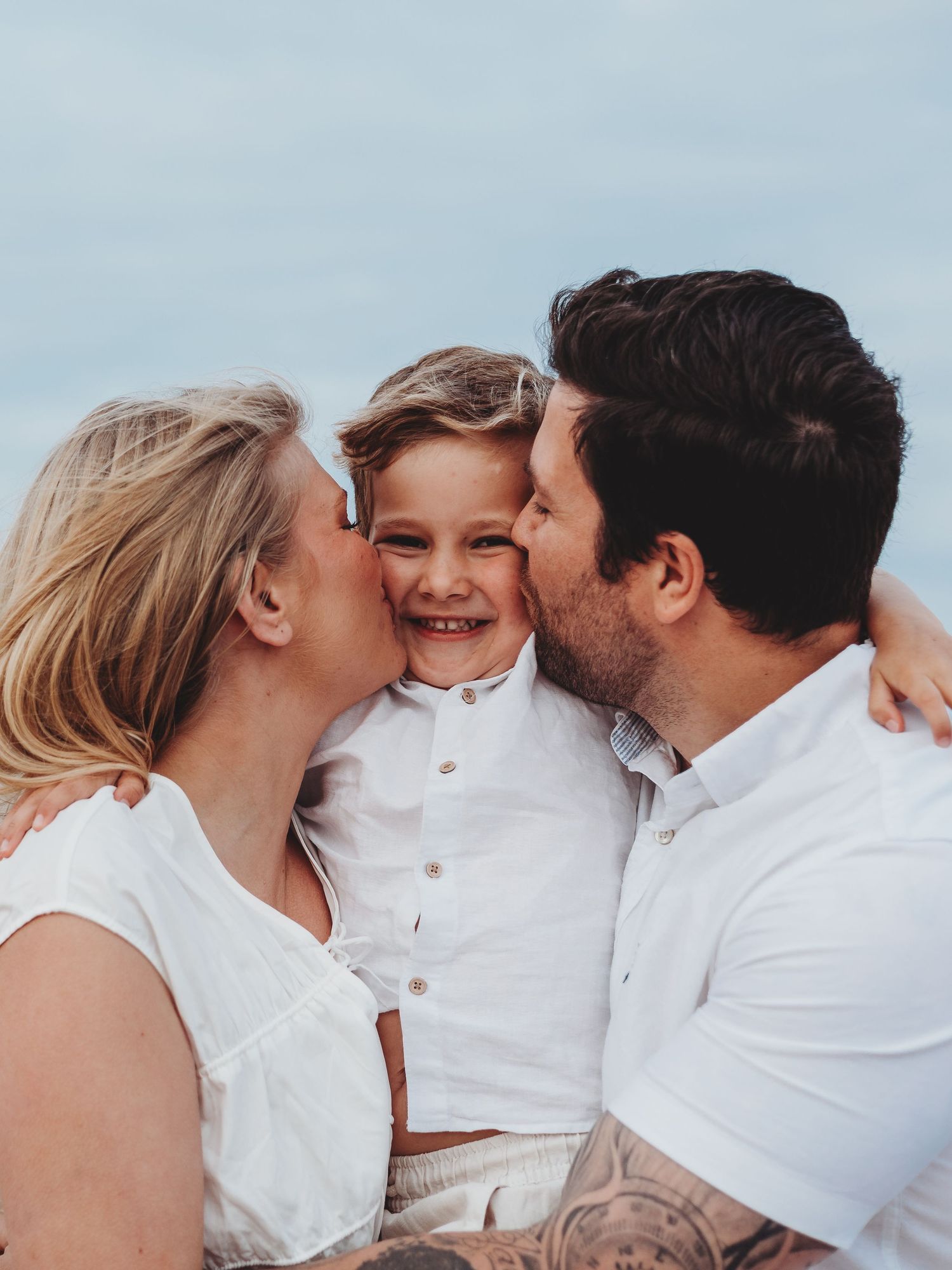 A family wearing white clothing shares intimate kisses and embraces in a series of tender outdoor portrait photographs.