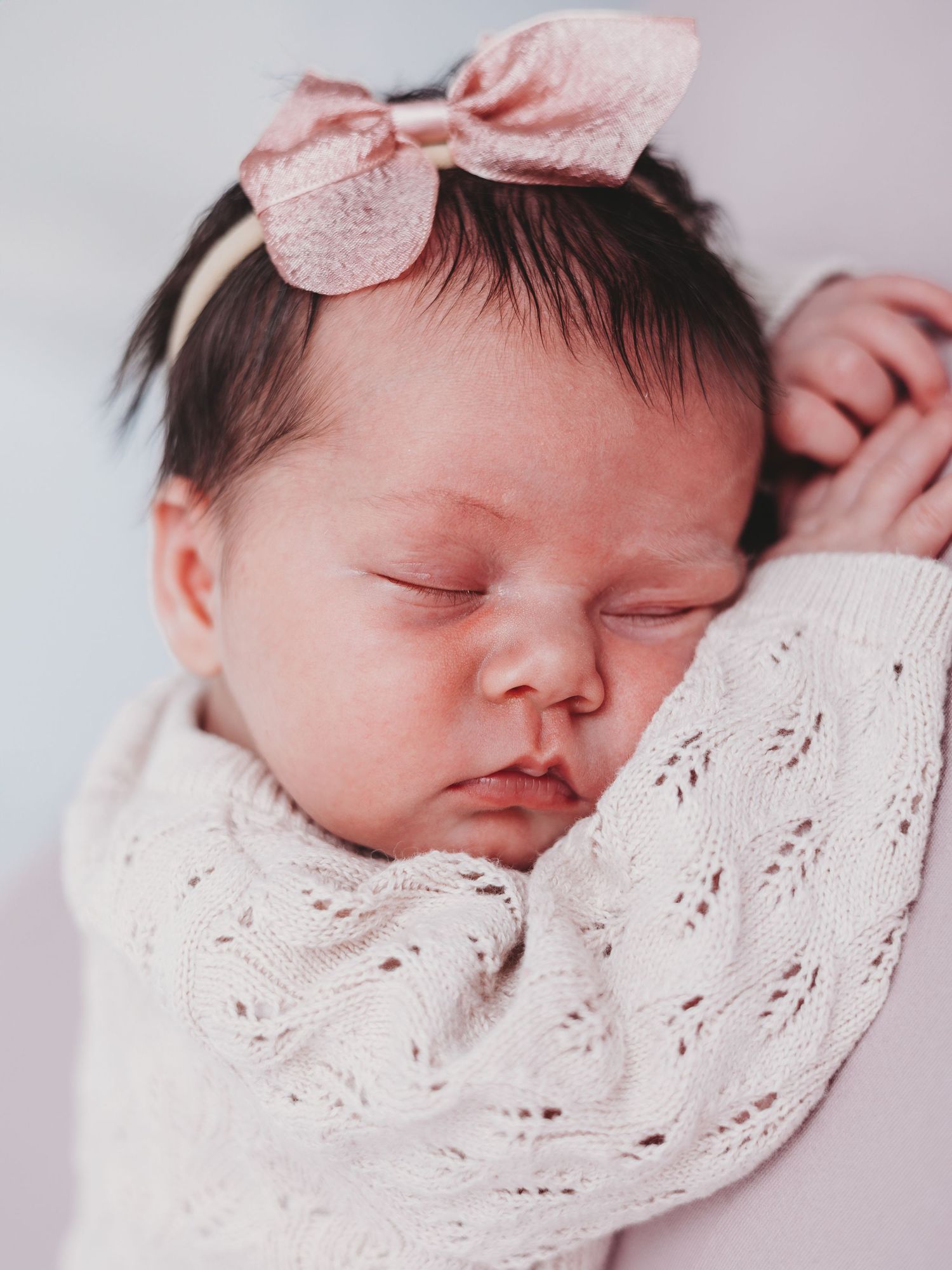 Sleeping newborn baby wearing a pink bow and white lace swaddle in soft lighting.