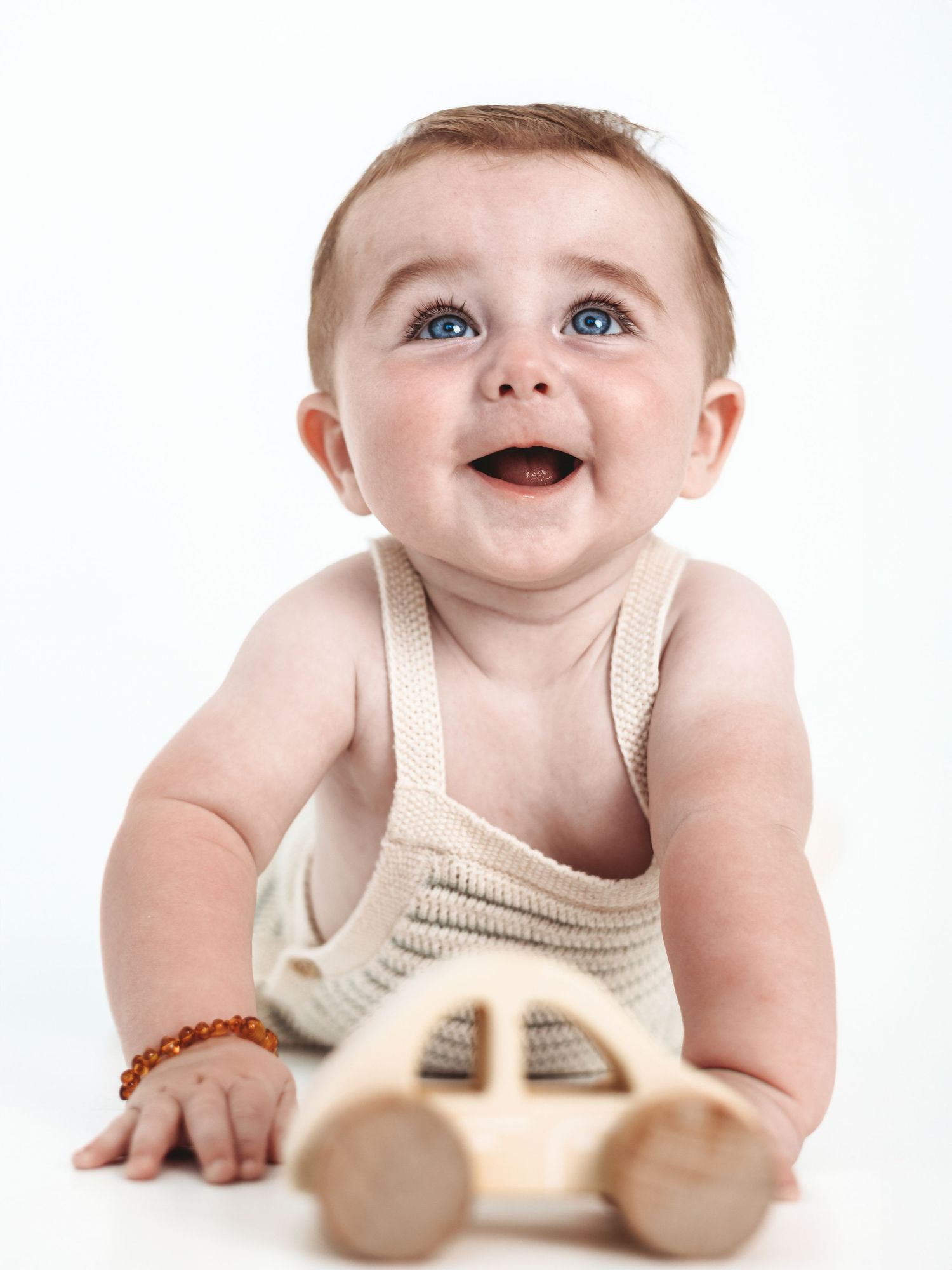 Smiling baby in white tank top playing with wooden toy car on white background.