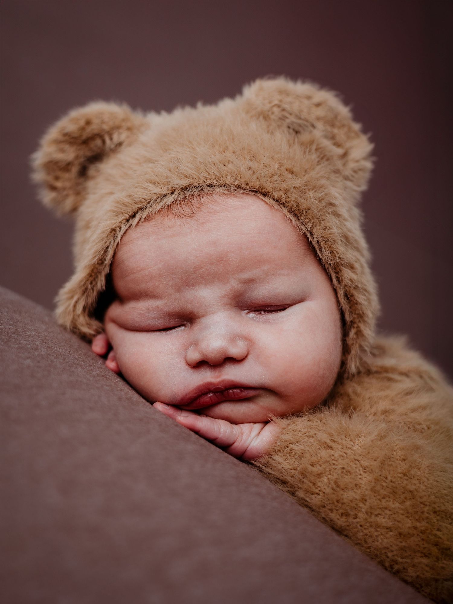A sleeping infant wearing a cozy brown bear costume with fuzzy ears lies peacefully against a dark background.
