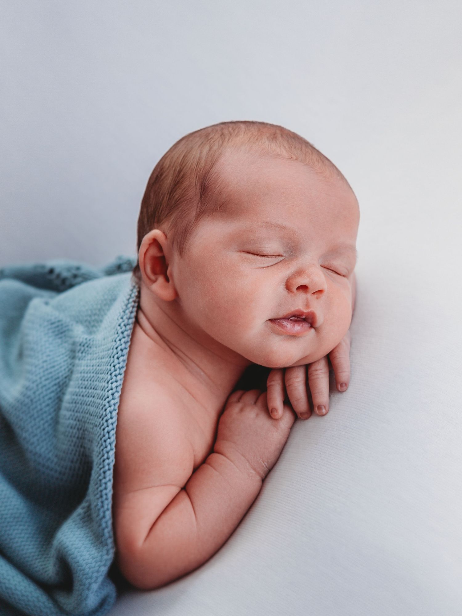 Peaceful sleeping newborn baby wrapped in soft blue blanket resting on white backdrop during professional photoshoot.