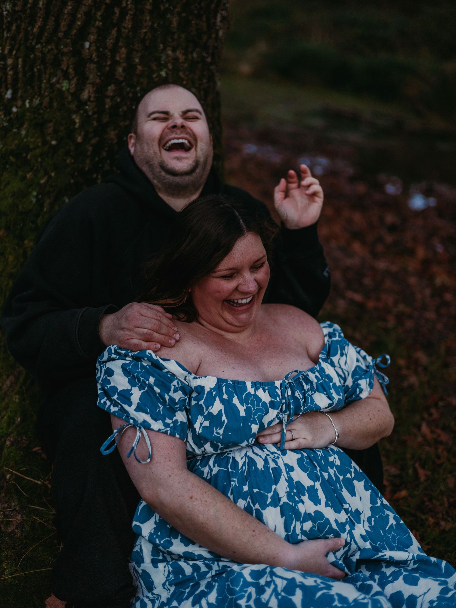 A couple shares a joyful moment of laughter while sitting against a tree trunk in an outdoor setting.