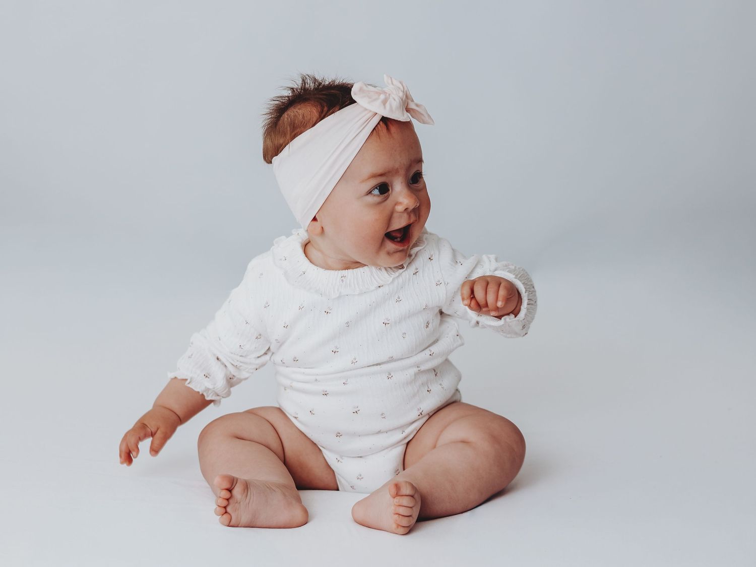 Adorable baby girl in white outfit and pink bow headband sitting and smiling on a light gray background.