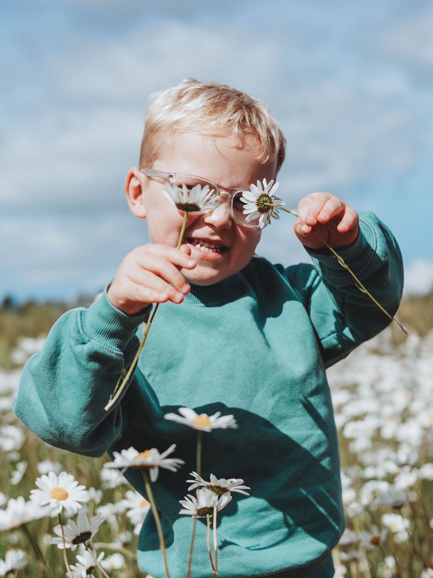 A young blonde boy in a teal sweatshirt and glasses holds daisies in a sunny flower field.