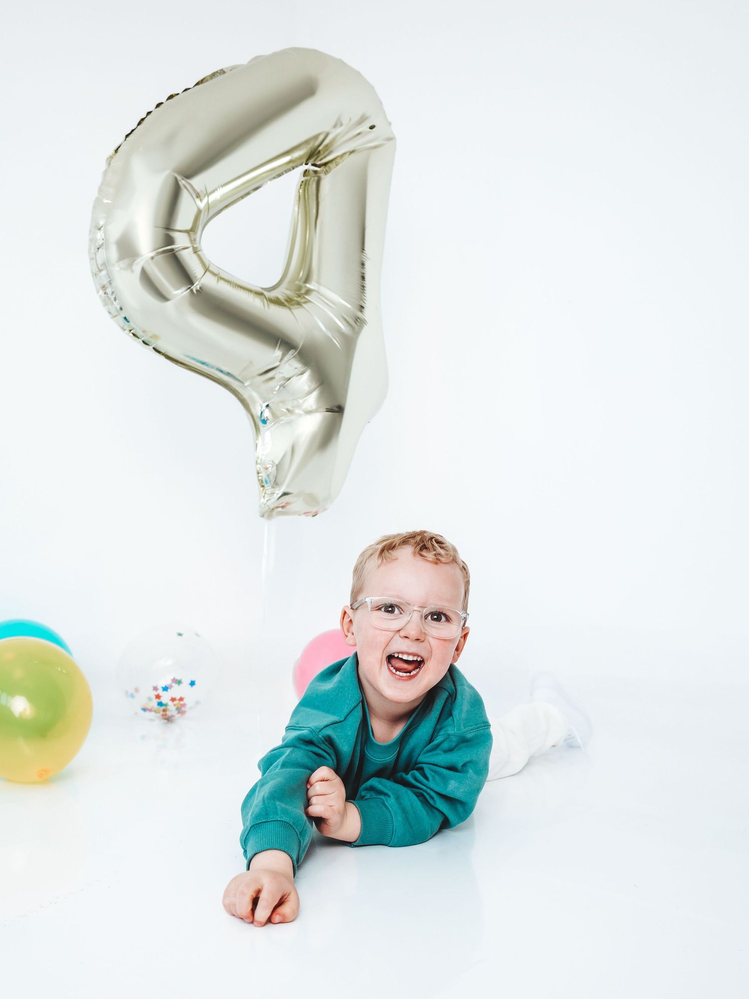 A happy toddler in teal outfit lies on white floor beside a large silver number 4 balloon and colorful balloons.