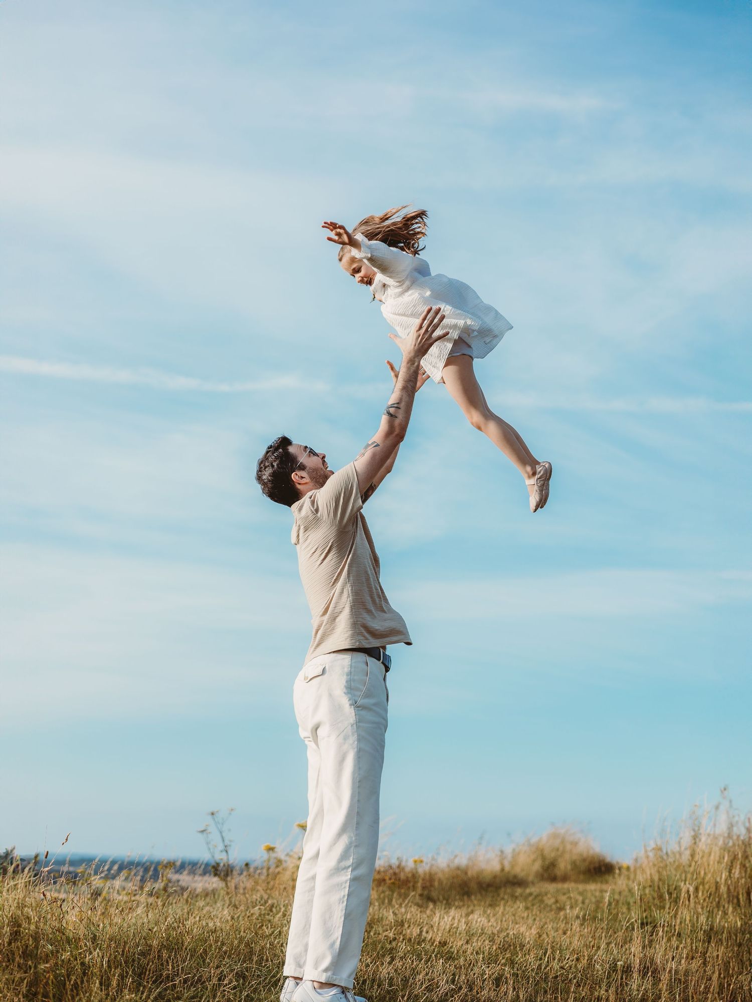 A father joyfully tosses his young daughter high into the air in a sunny field with a blue sky backdrop.