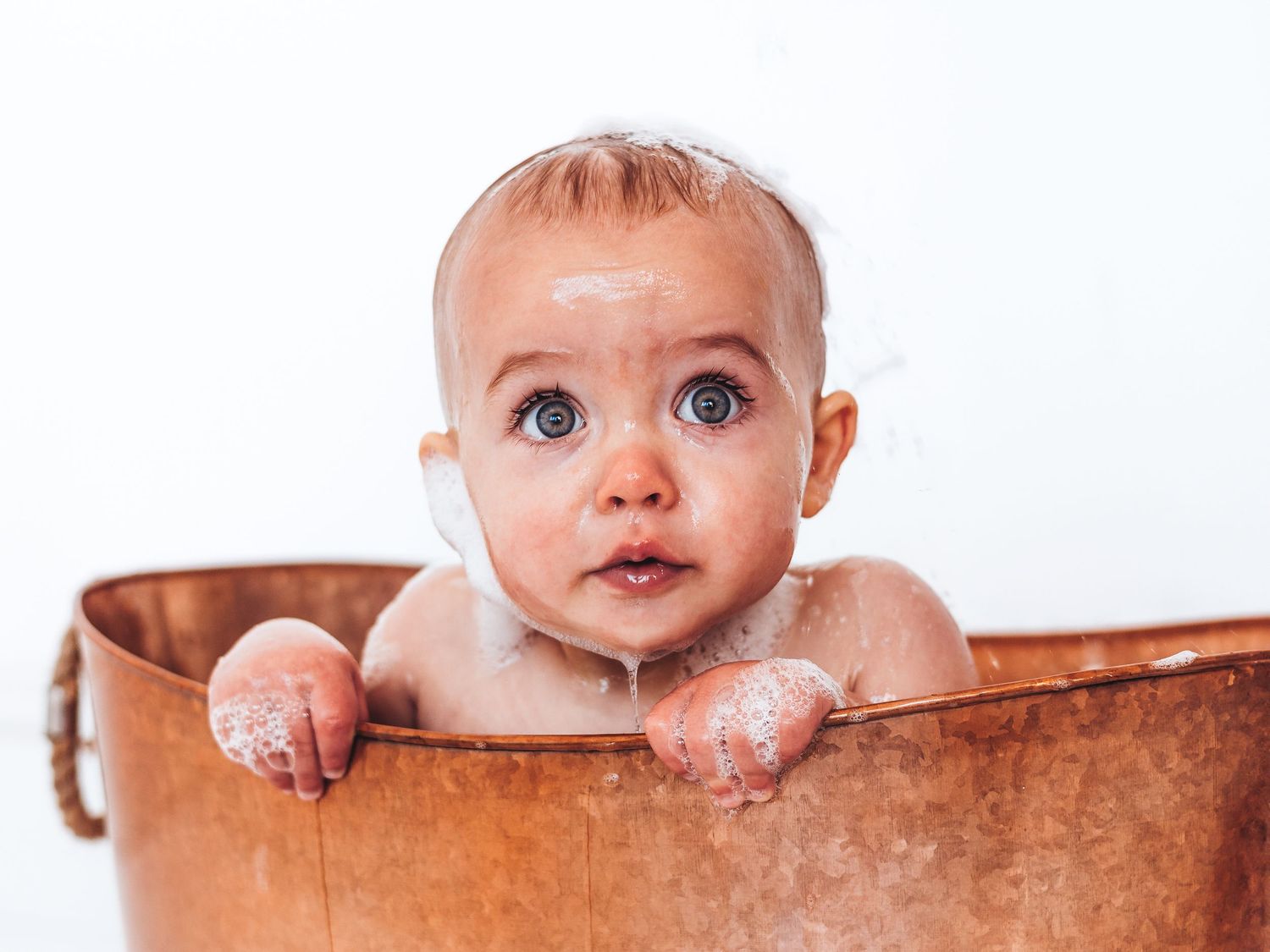 Adorable baby with wide blue eyes sitting in a rustic copper bathtub covered in soap suds.