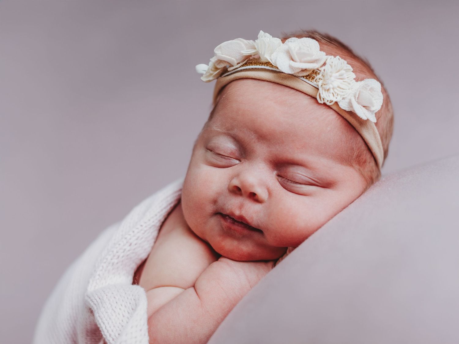 A peaceful sleeping newborn baby wearing a delicate white headband and wrapped in soft fabric.
