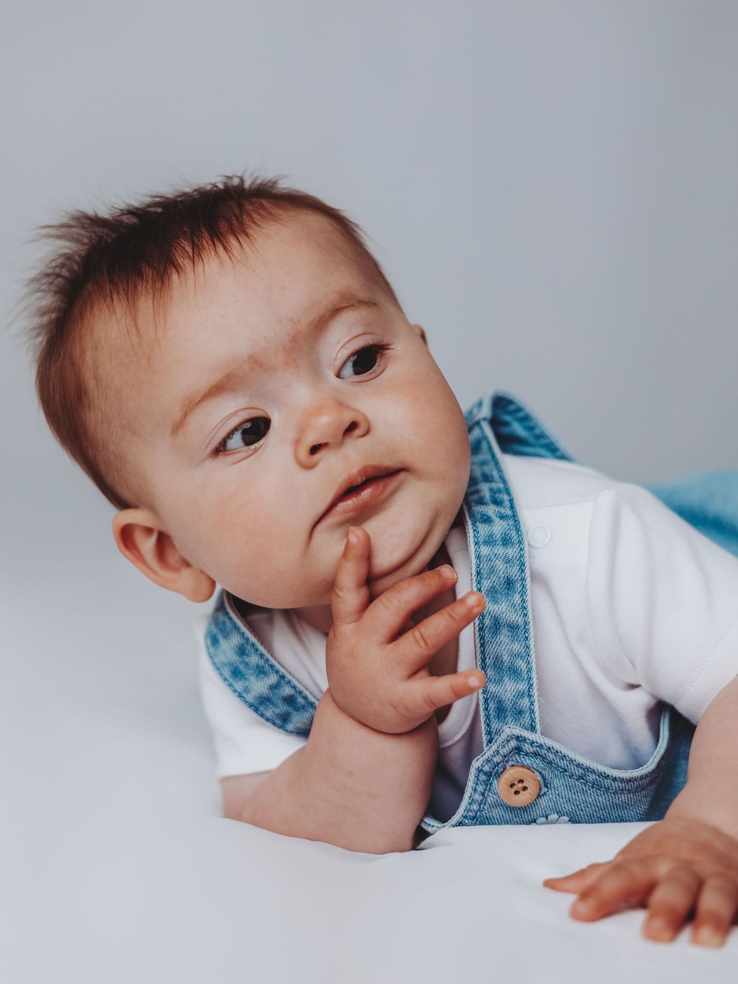 Adorable baby in denim overalls and white shirt lying down with finger near mouth in thoughtful expression.