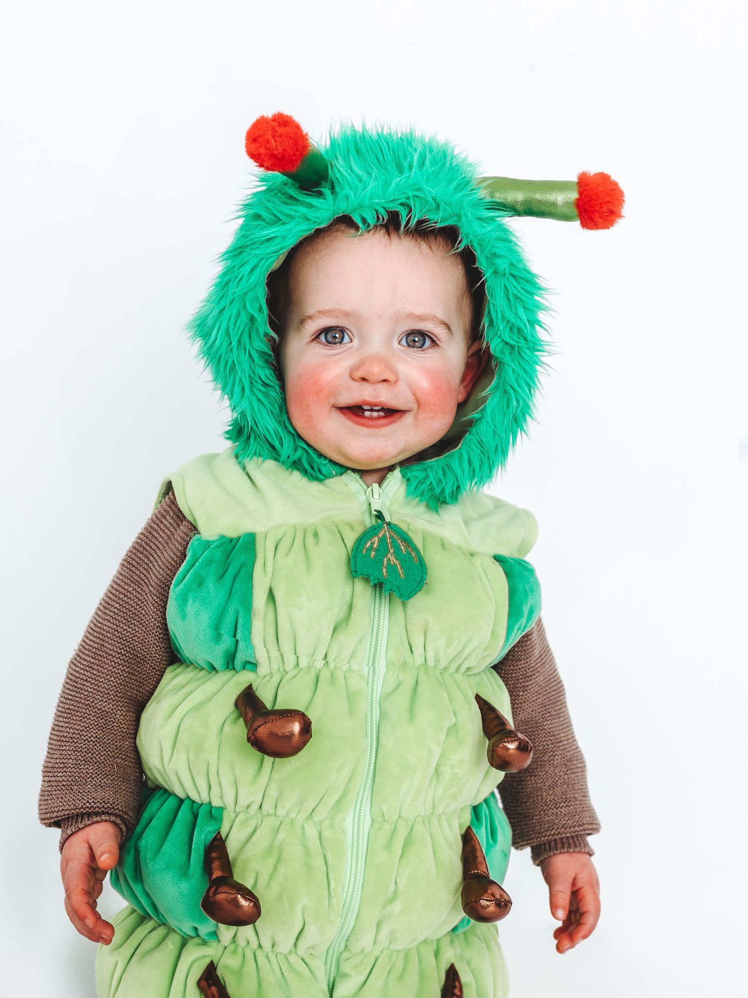 Smiling baby wearing a green caterpillar Halloween costume with fuzzy hood and red antennae against white background.
