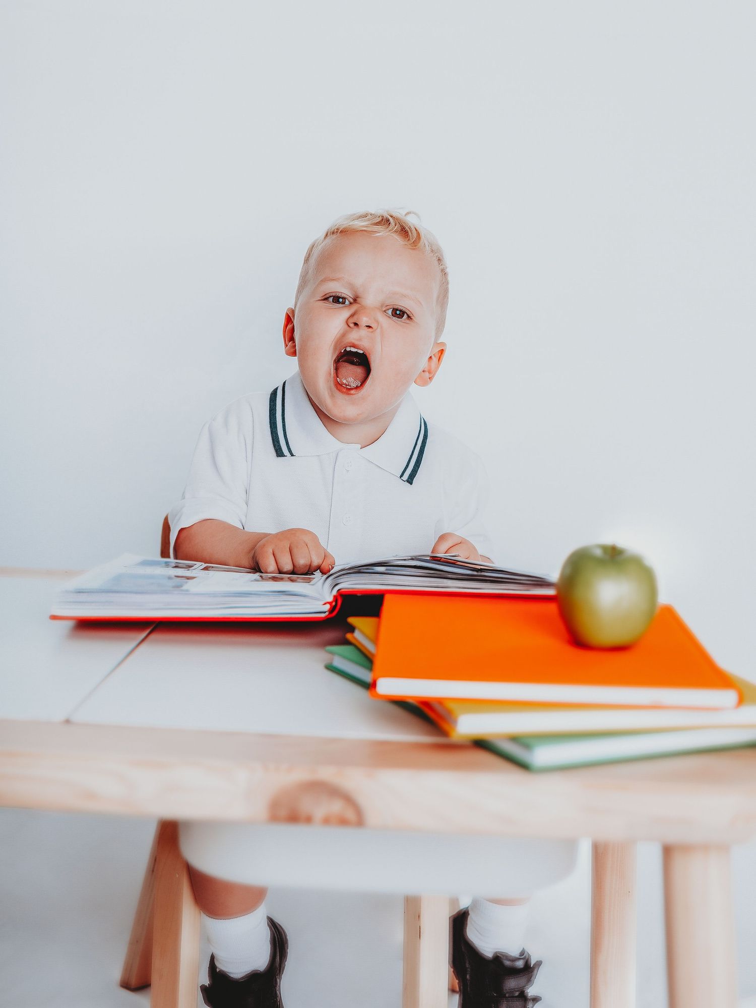Toddler boy sitting at wooden desk with open book, green apple, and colorful notebooks, mouth open in surprise.
