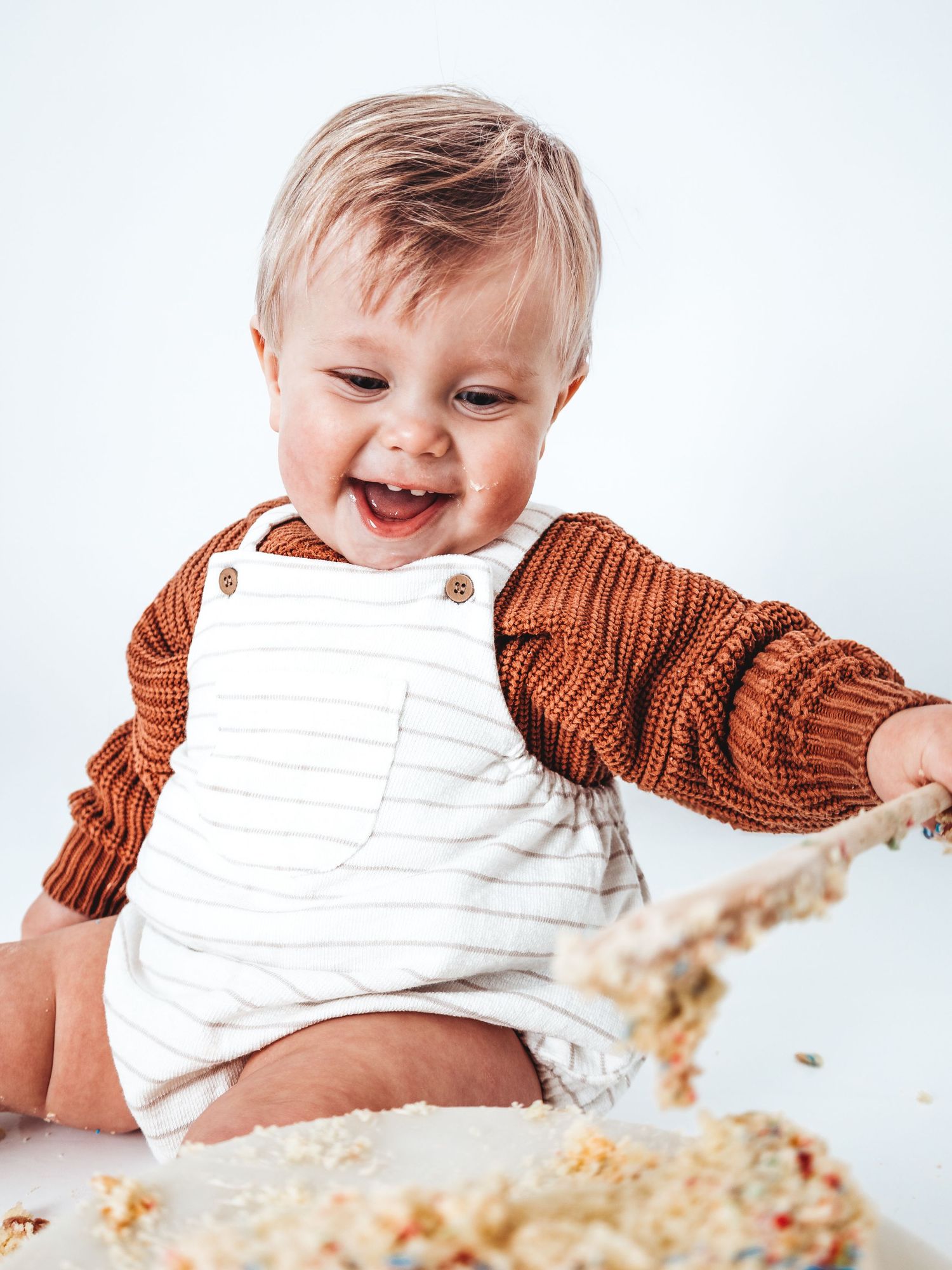 Happy baby in striped overalls and rust knit sweater smashing birthday cake, laughing with joy during first birthday celebration.