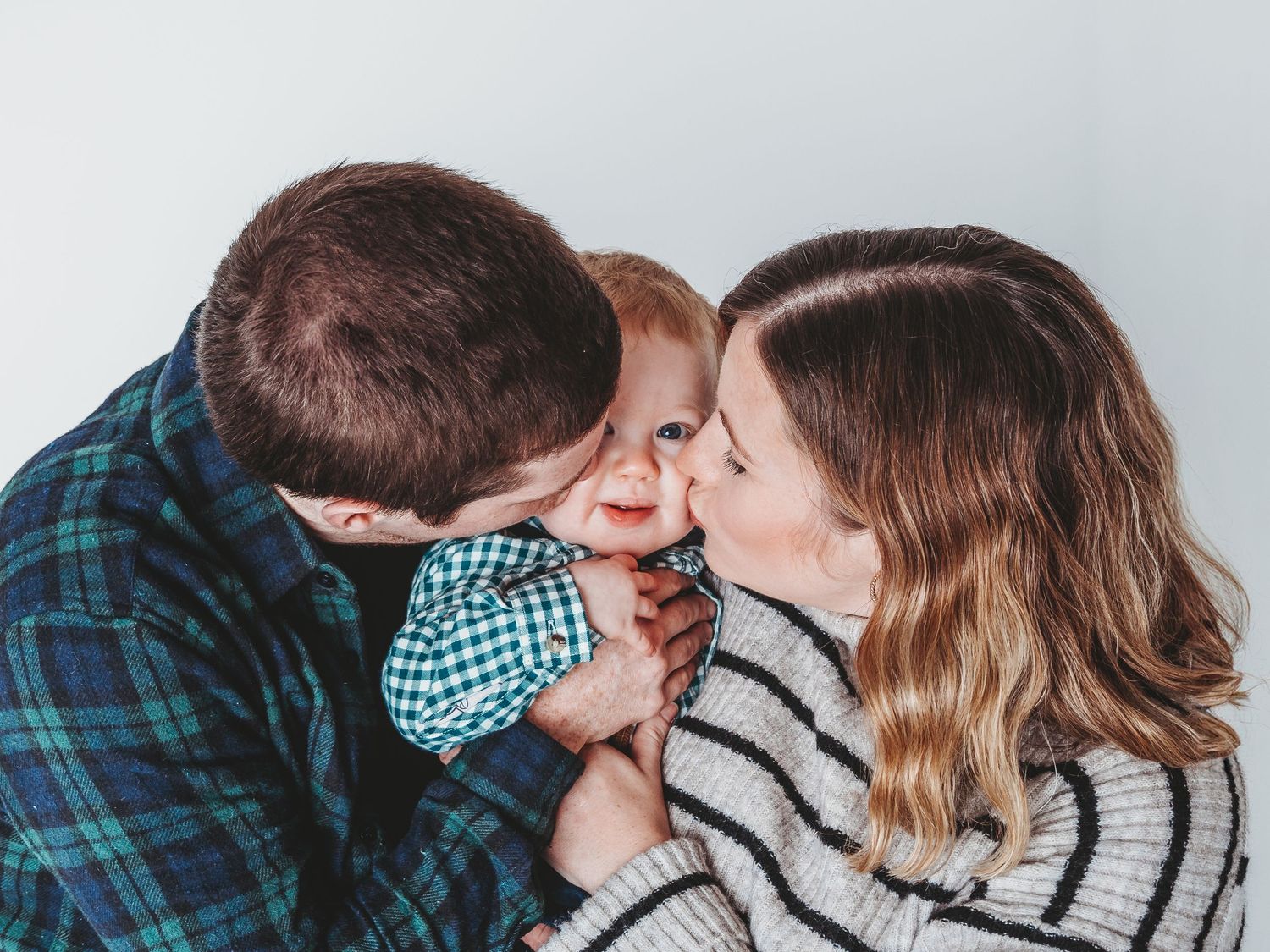 Two parents lovingly kiss their smiling baby on the cheeks in a heartwarming family portrait.