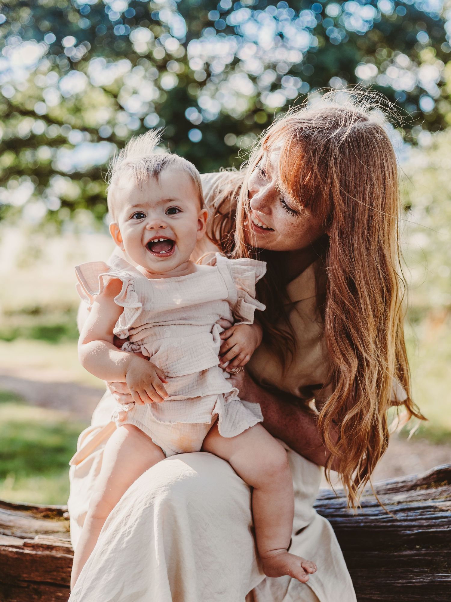 A joyful baby in a ruffled outfit laughs while being held by a smiling woman with long hair outdoors.