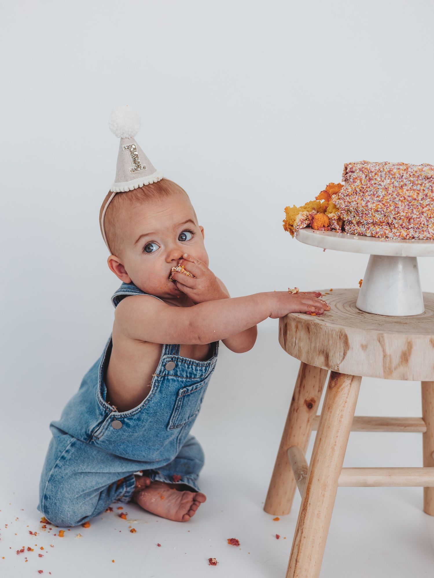 A baby in denim overalls reaches for a white cake on a wooden stool in a progression of playful photos.