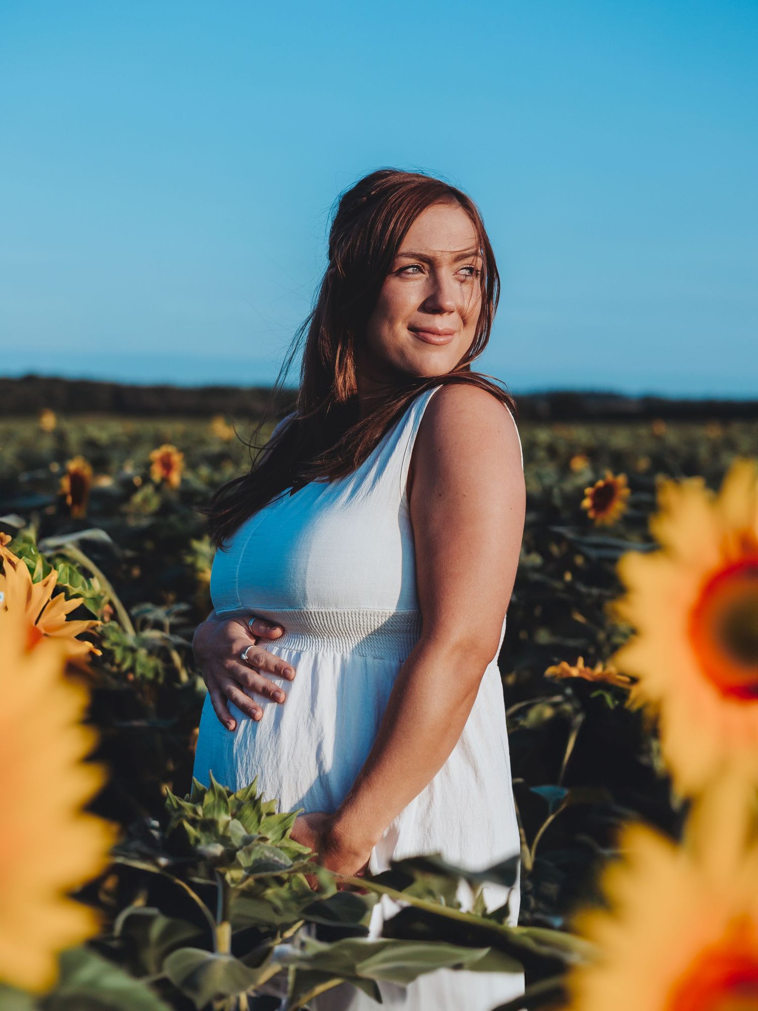 A person in a white dress poses gracefully among blooming sunflowers in a field during golden hour.