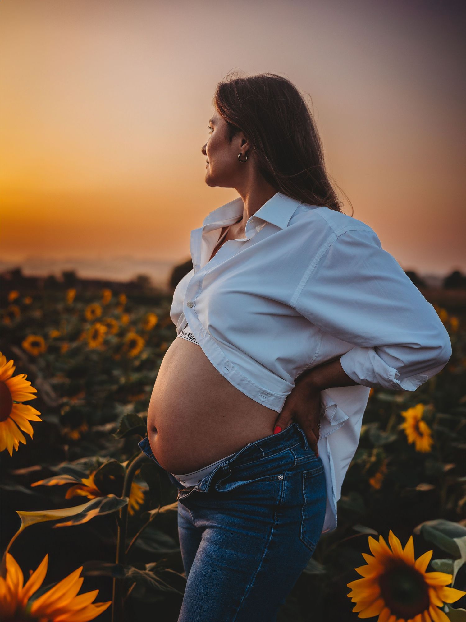 Maternity photo series showing silhouette against sunset in sunflower field with white shirt and jeans.