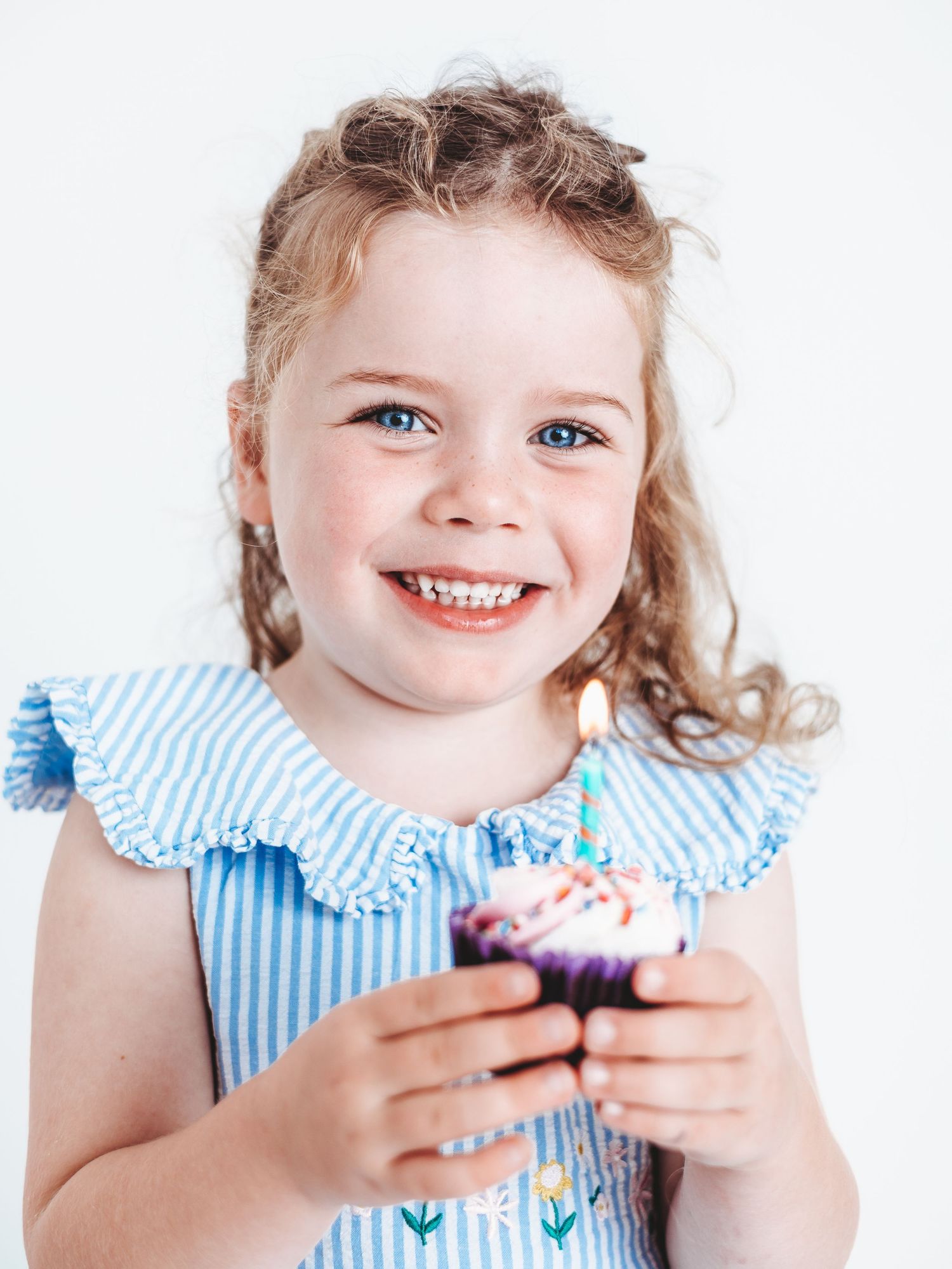 A child in a blue striped dress holds a cupcake with pink frosting and sprinkles, smiling with joy.