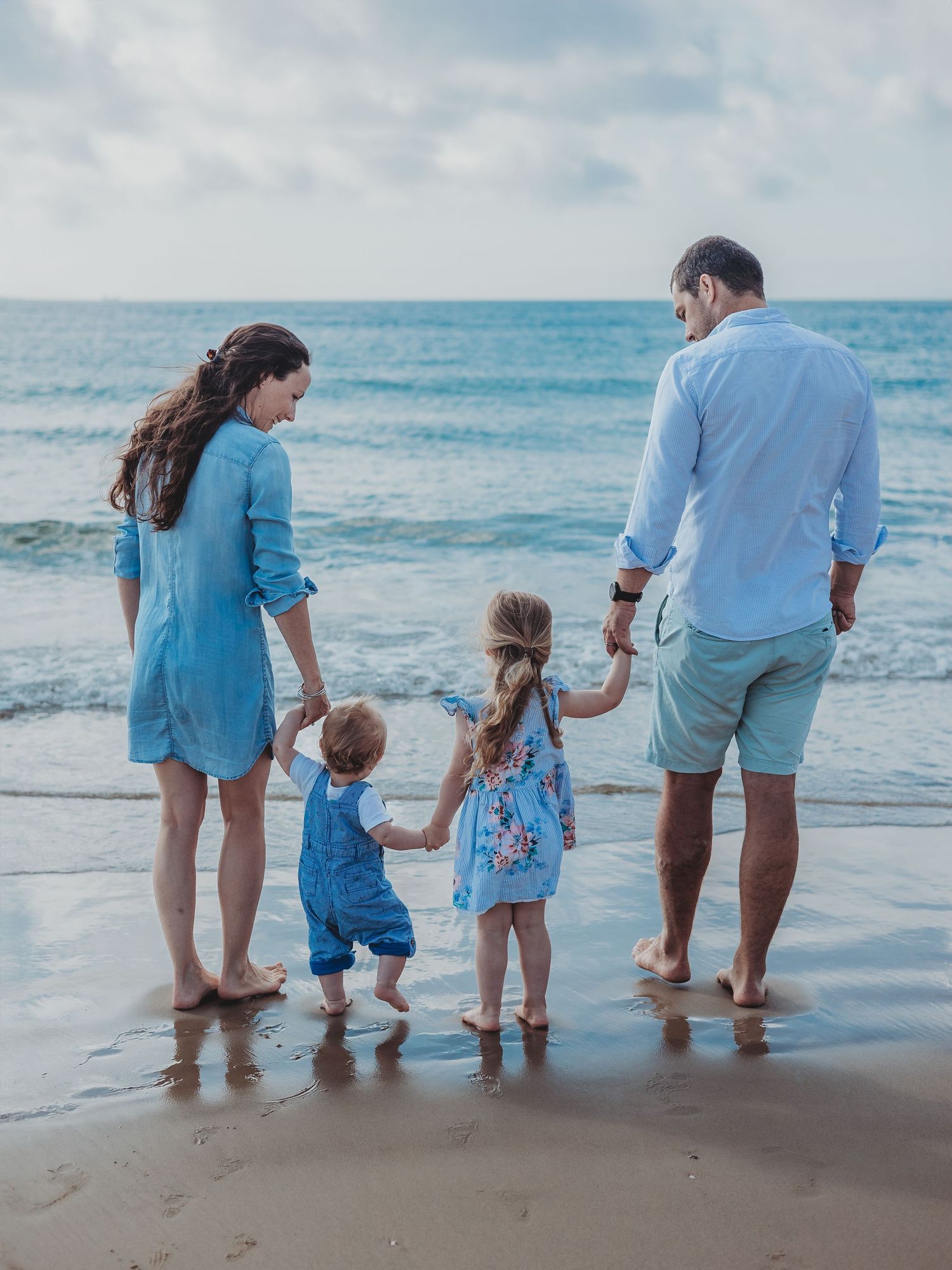 Family of four holding hands on a sandy beach shore, parents with two young children facing the ocean waves.