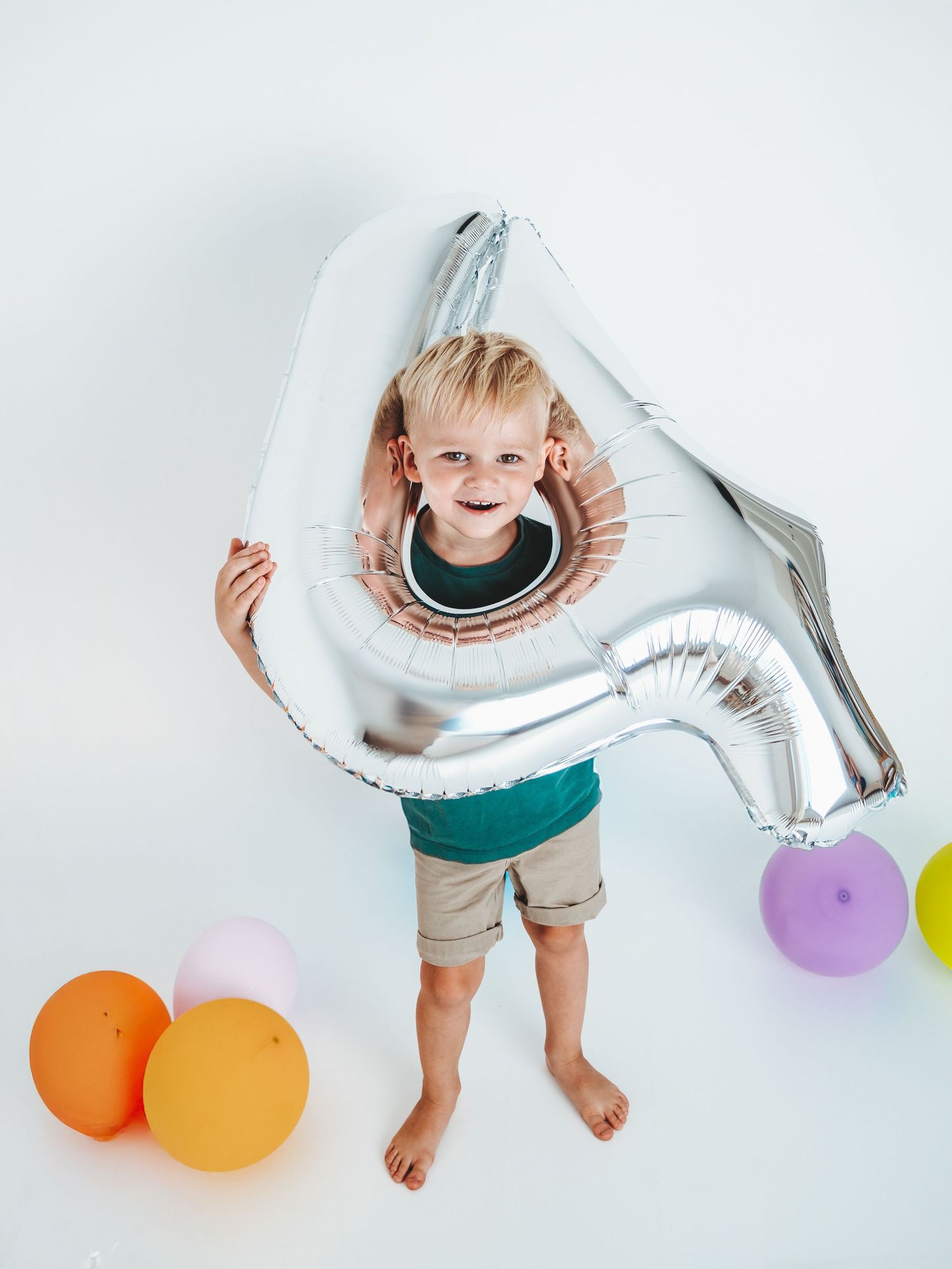 Smiling blonde toddler holding a large silver number balloon surrounded by colorful balloons on white background.
