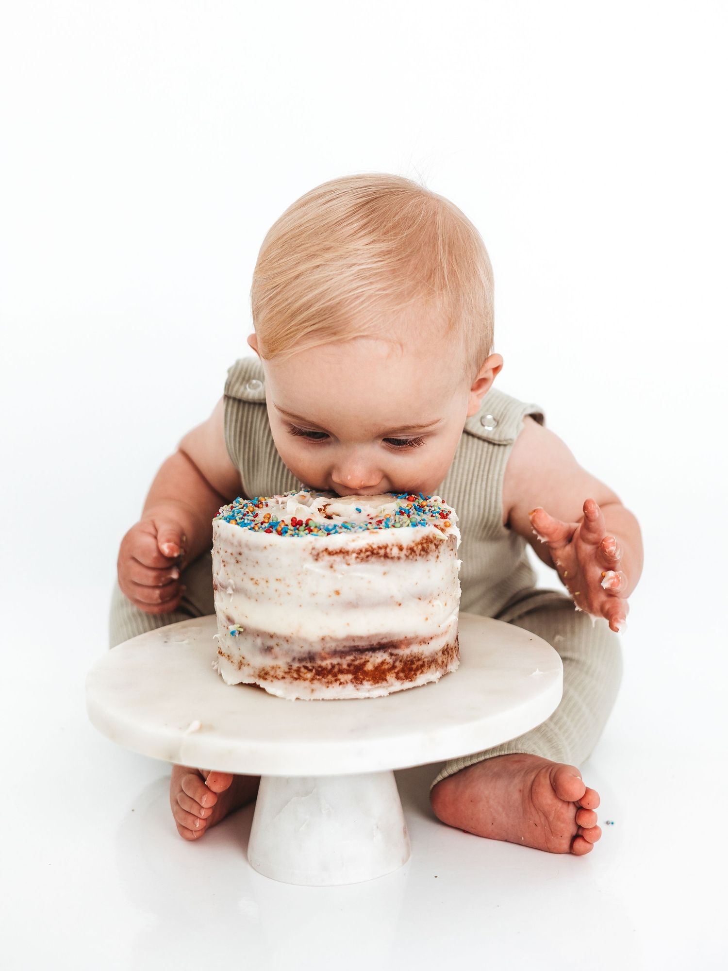 Baby in green overalls smashing face into first birthday cake with colorful sprinkles on white cake stand.