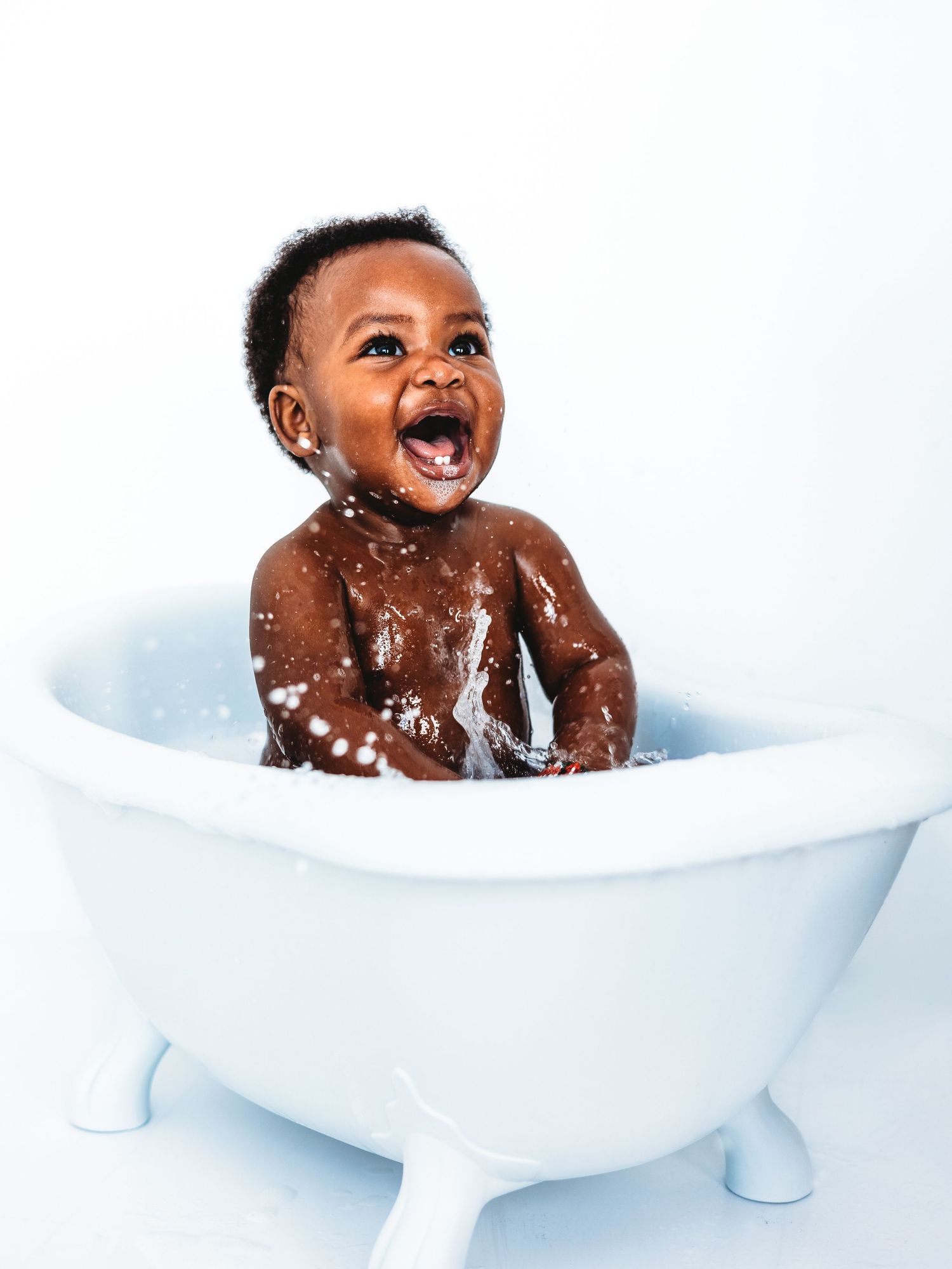 Joyful Black baby laughing and splashing in a small white clawfoot bathtub against a white background.