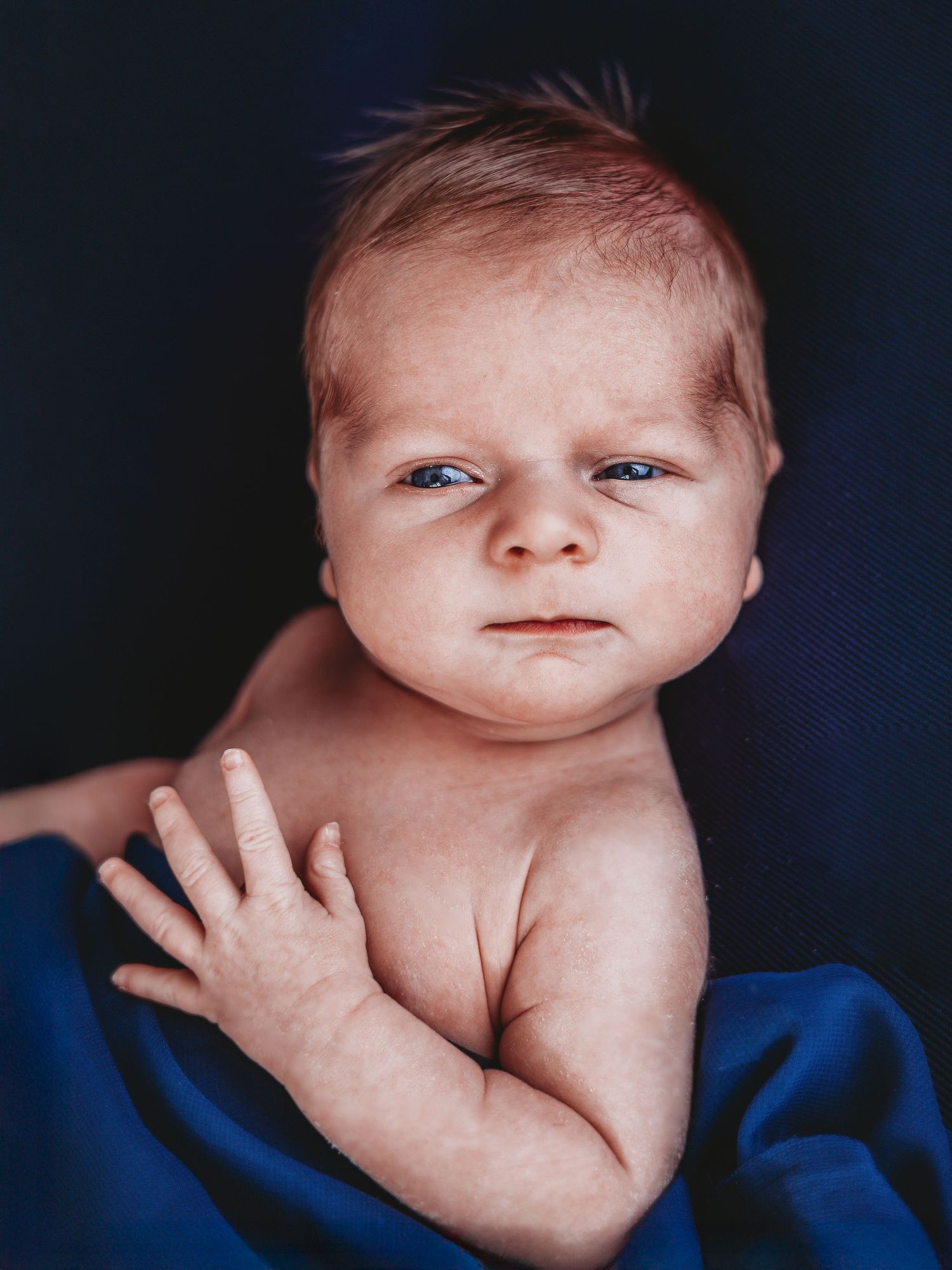 A newborn baby with bright eyes lies on blue fabric while raising one hand in a portrait-style photograph.