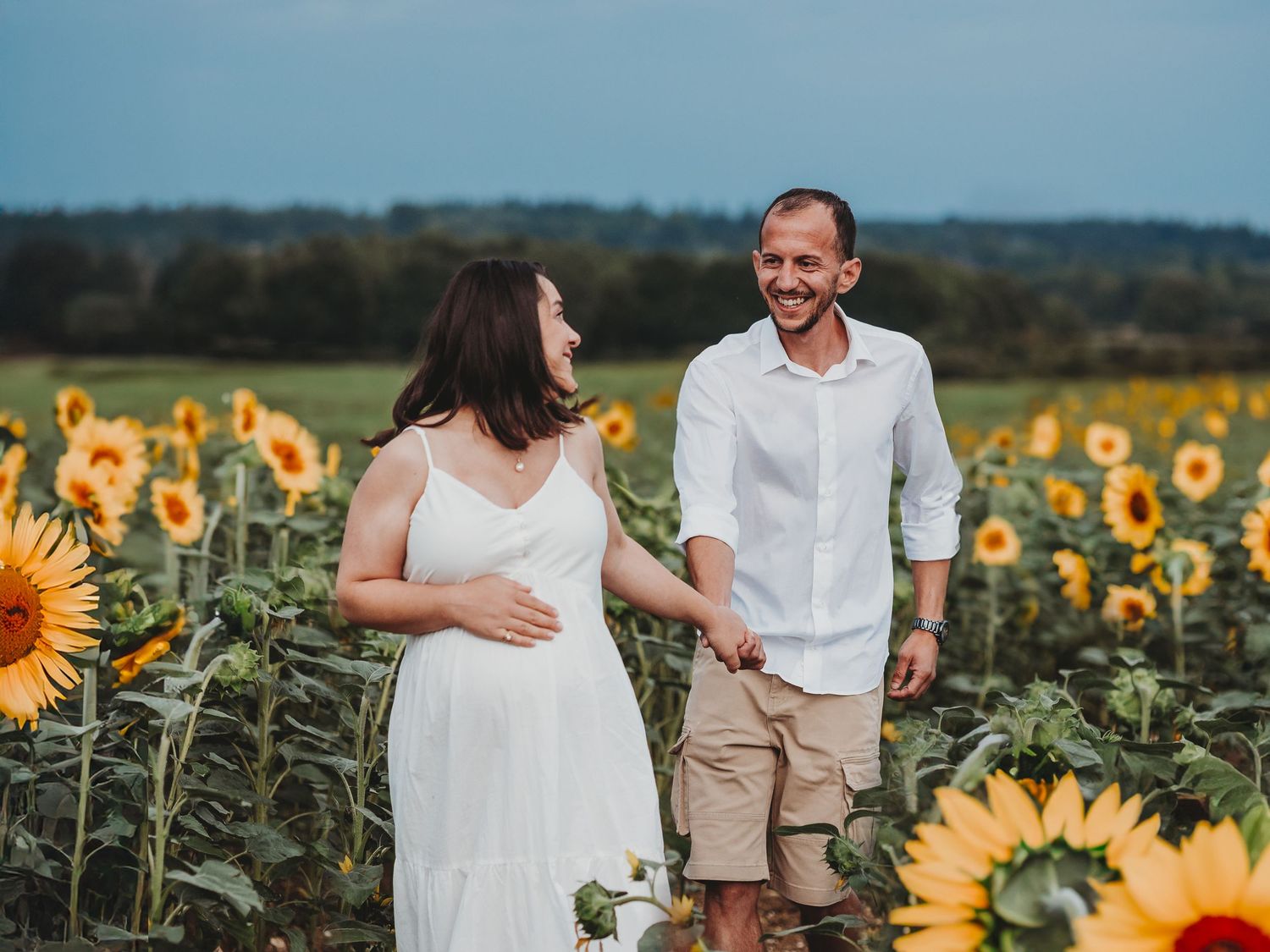 Couple walks hand in hand through vibrant yellow sunflower field on sunny summer day.