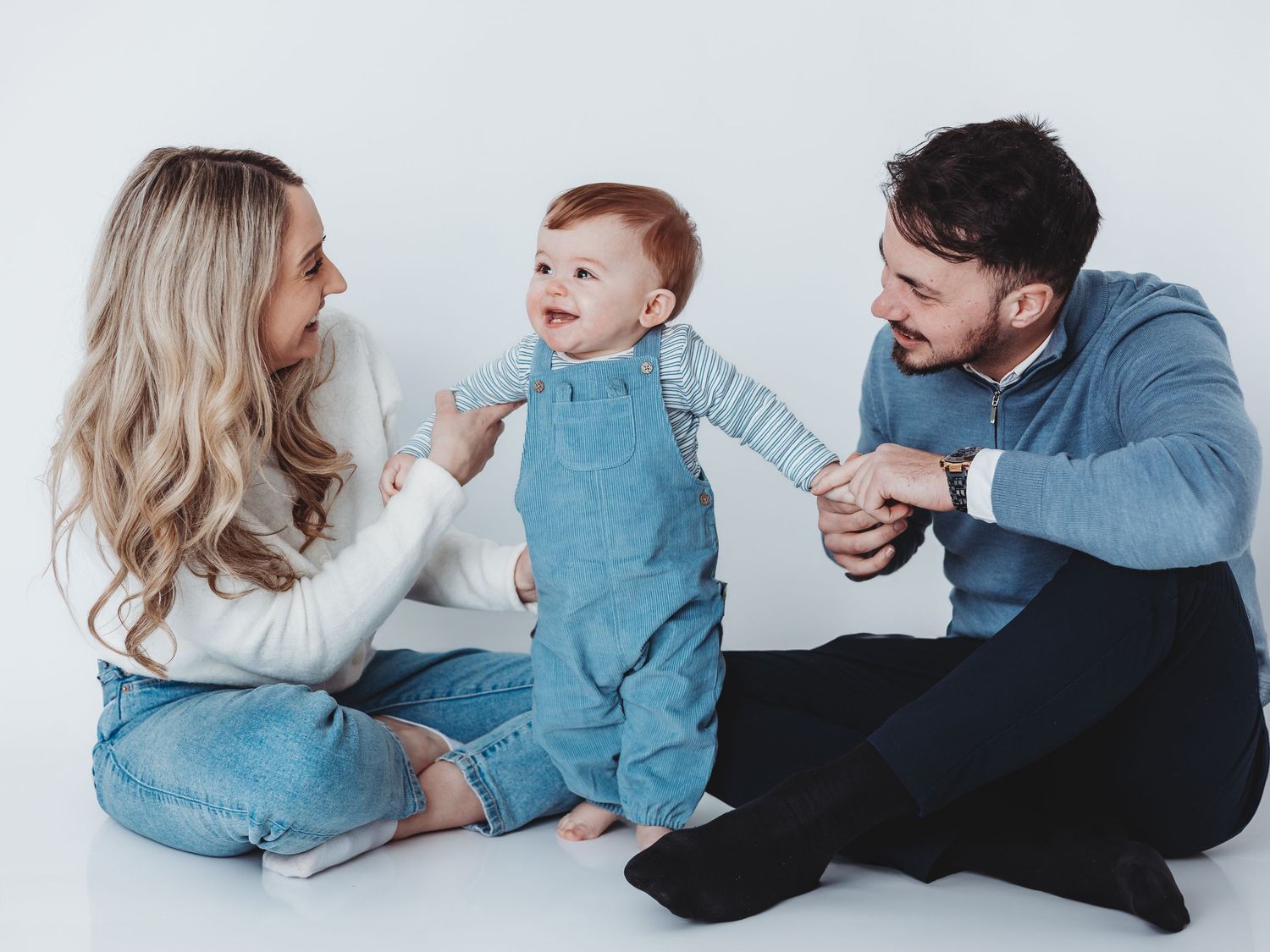 Happy young family with a red-haired baby sitting on a white studio floor, smiling and playing together.
