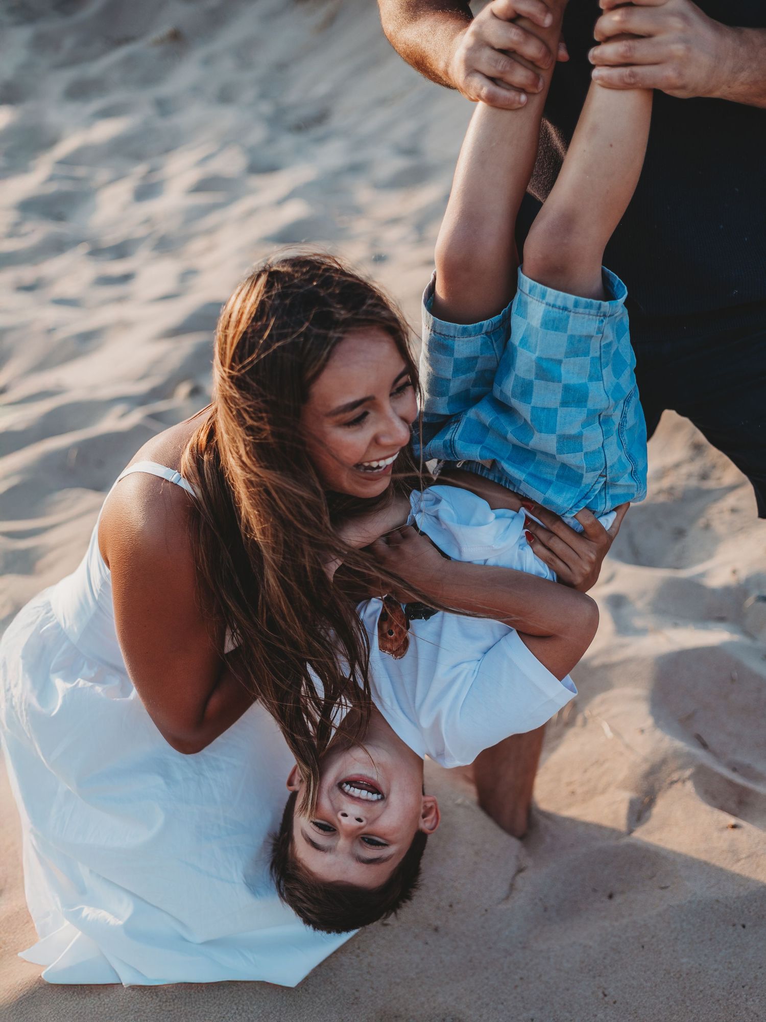 A smiling woman in a white dress plays with a laughing child held upside down on a sandy beach.