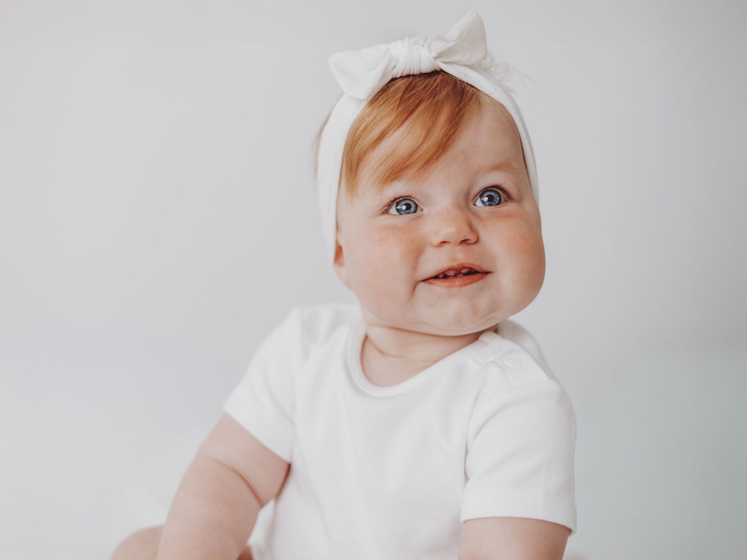 A smiling baby wearing a white t-shirt and white bow headband against a light gray background.