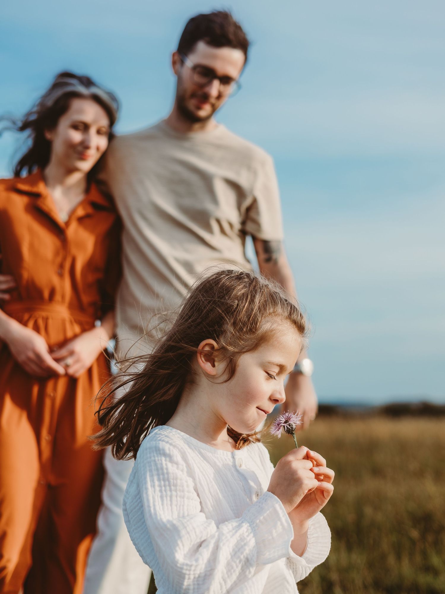 A young girl smells a small purple flower in a sunny field while her parents smile warmly behind her.
