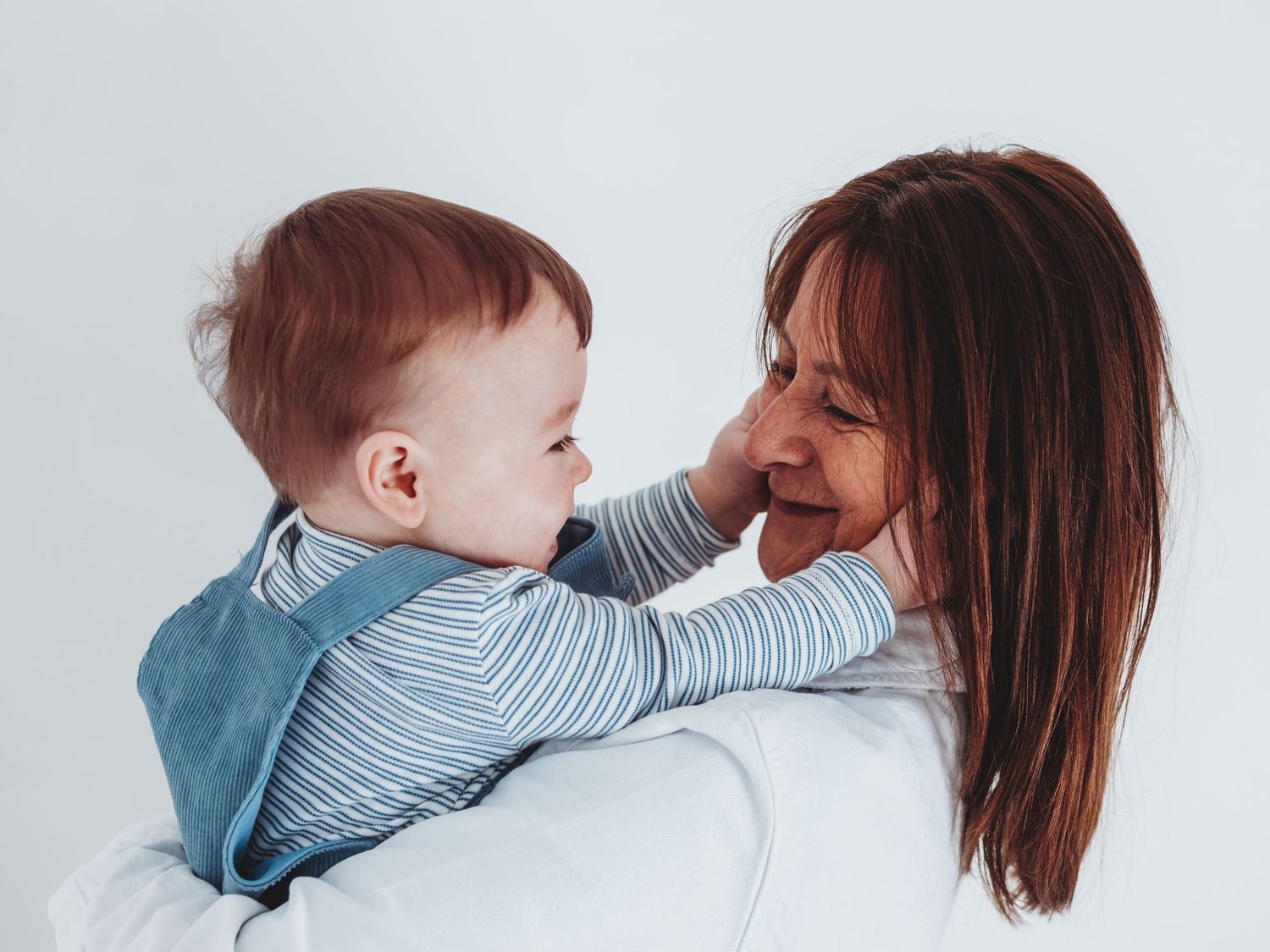 A joyful mother holds her redheaded baby who reaches up to touch her smiling face against a white background.