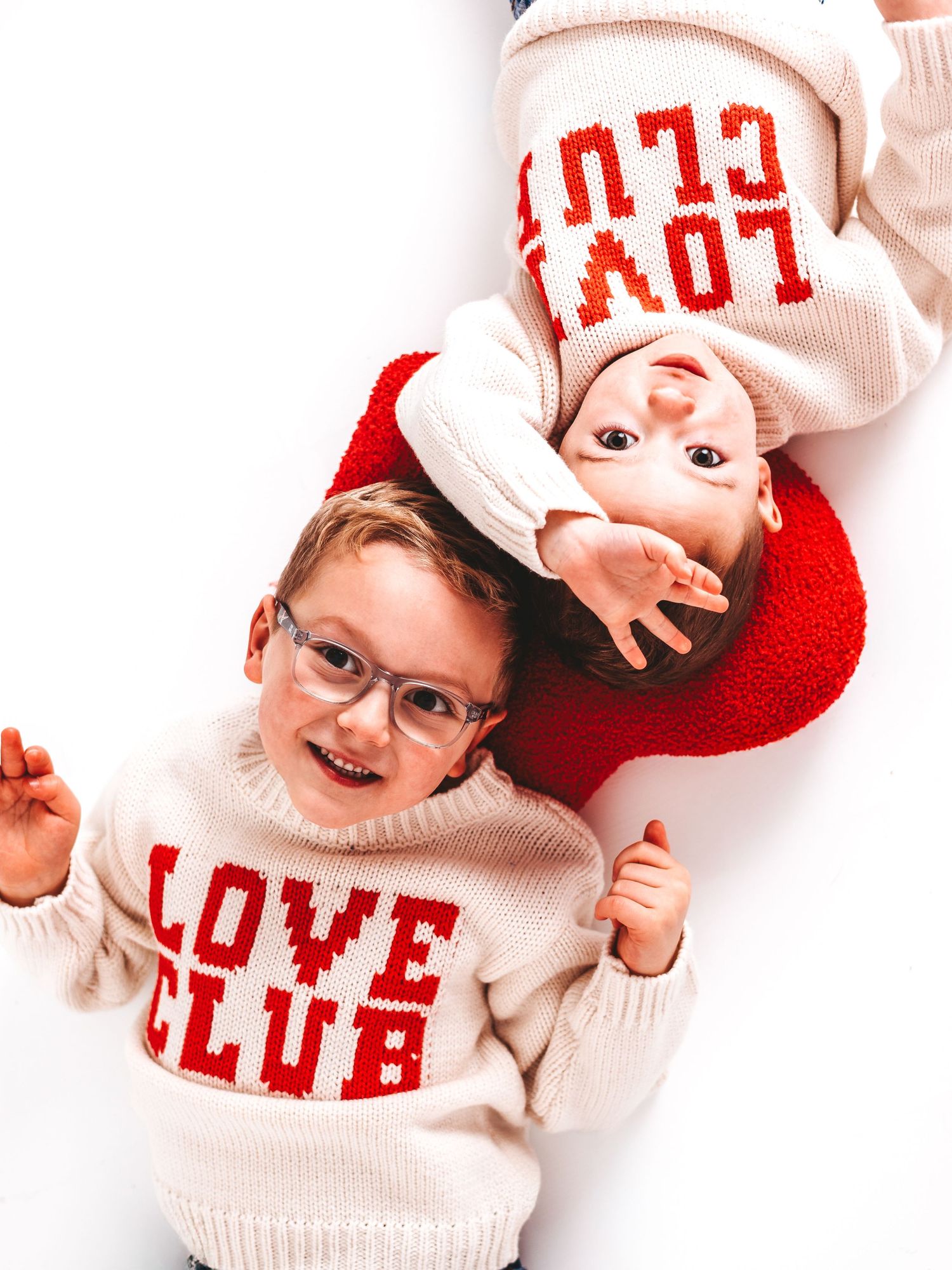 Two children wearing matching cream and red Love Club knit sweaters lying on a white background.