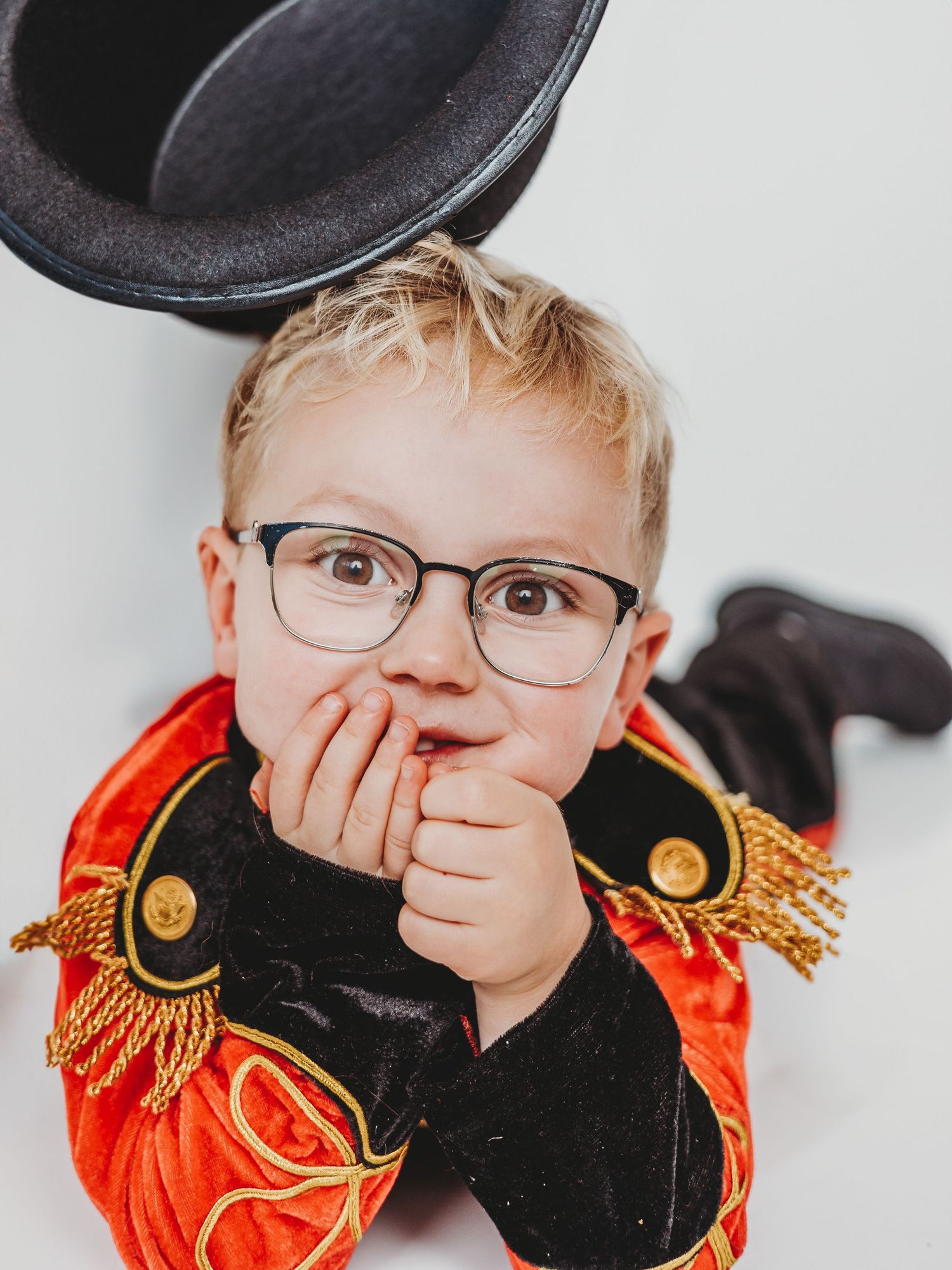Adorable toddler in ringmaster Halloween costume with glasses and top hat, lying down with hands on face.