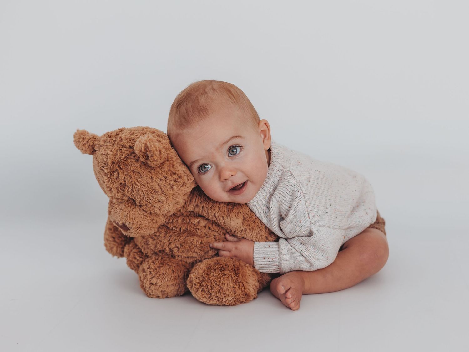 A baby in gray onesie lies on white bedding next to a brown teddy bear in a sweet portrait series.