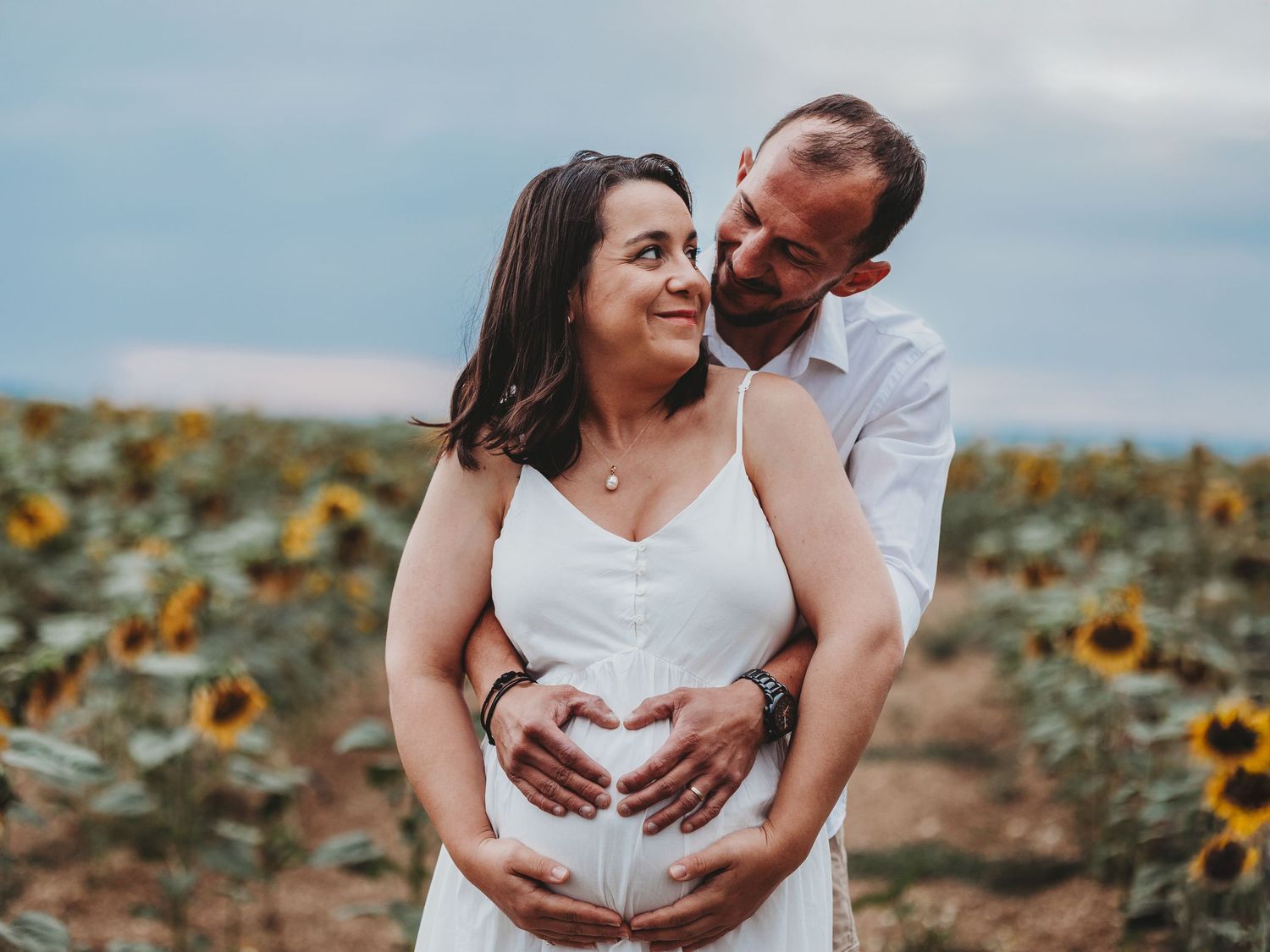 A couple embraces in a sunflower field during a maternity photo session at sunset.