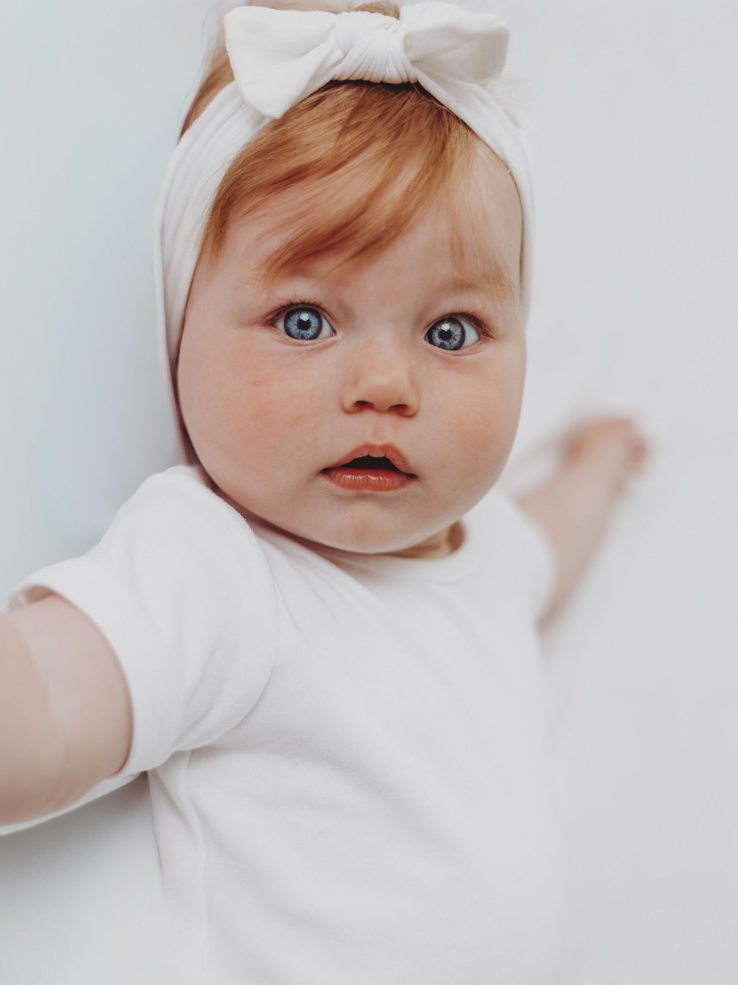 Baby wearing a white bow headband and white outfit lies against light background with bright blue eyes.