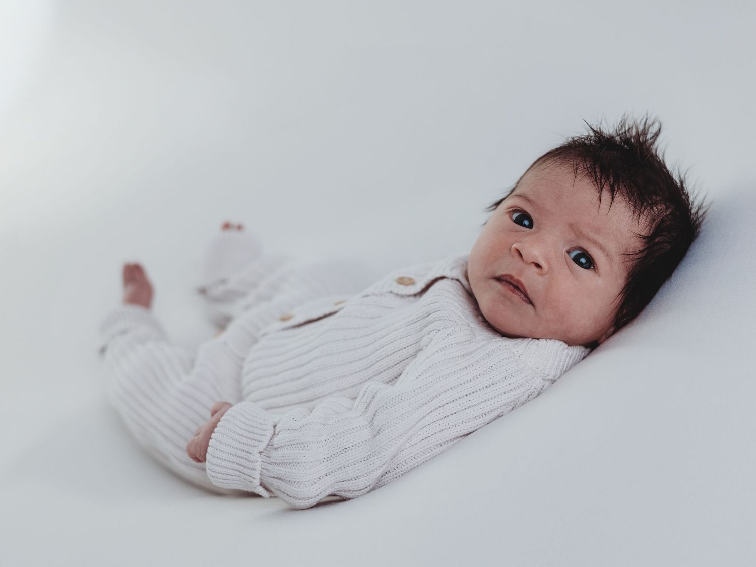 A baby wearing a white knit sweater lies on a white background looking to the side.